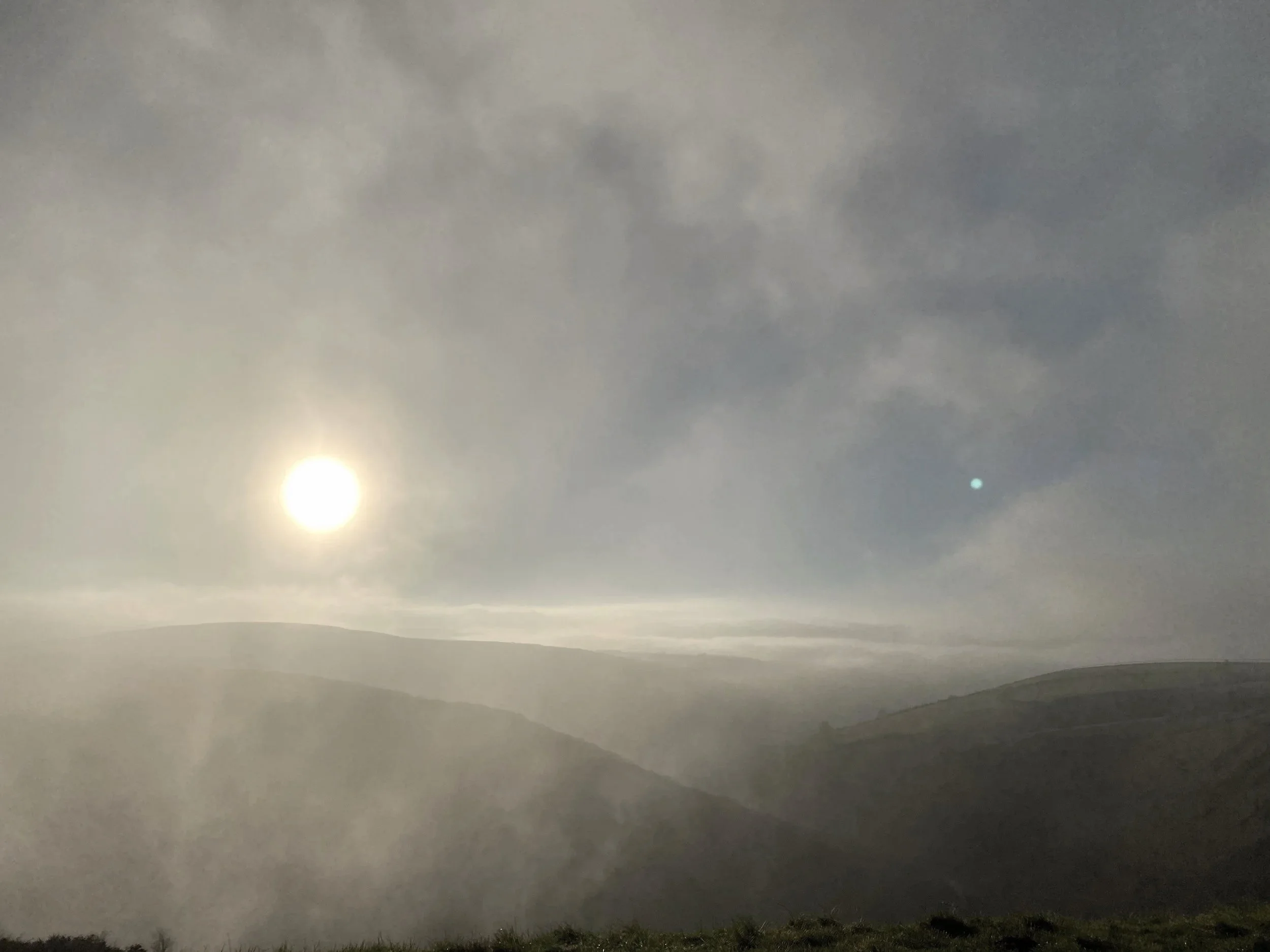 Landscape of hills with fog, the sun shining through clouds in a misty sky.
