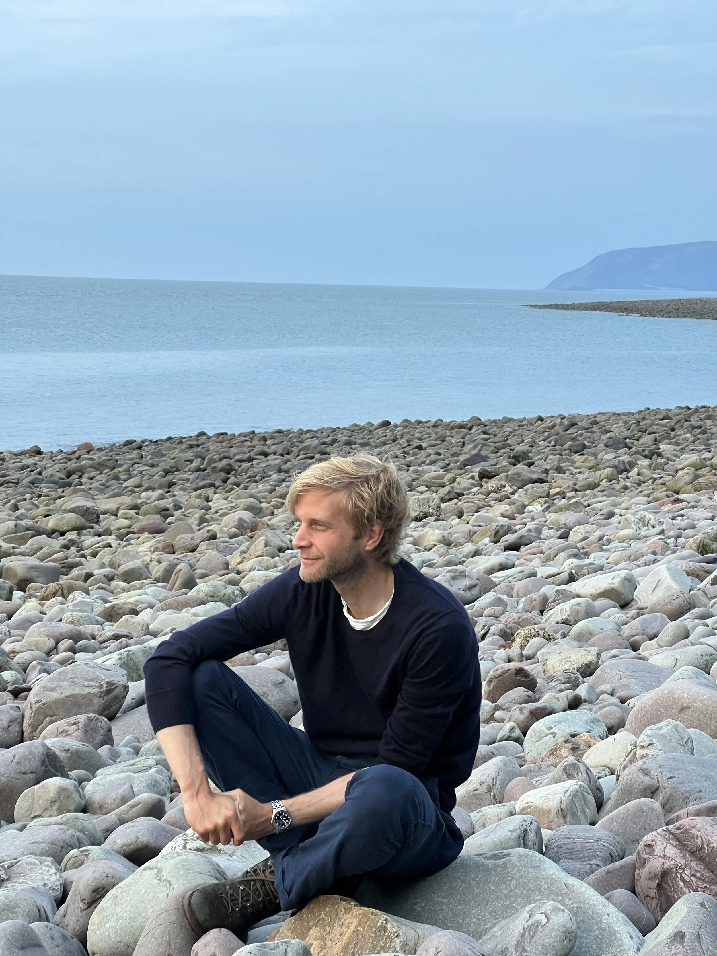 A man sitting on a rocky beach near the ocean, with cliffs in the background.