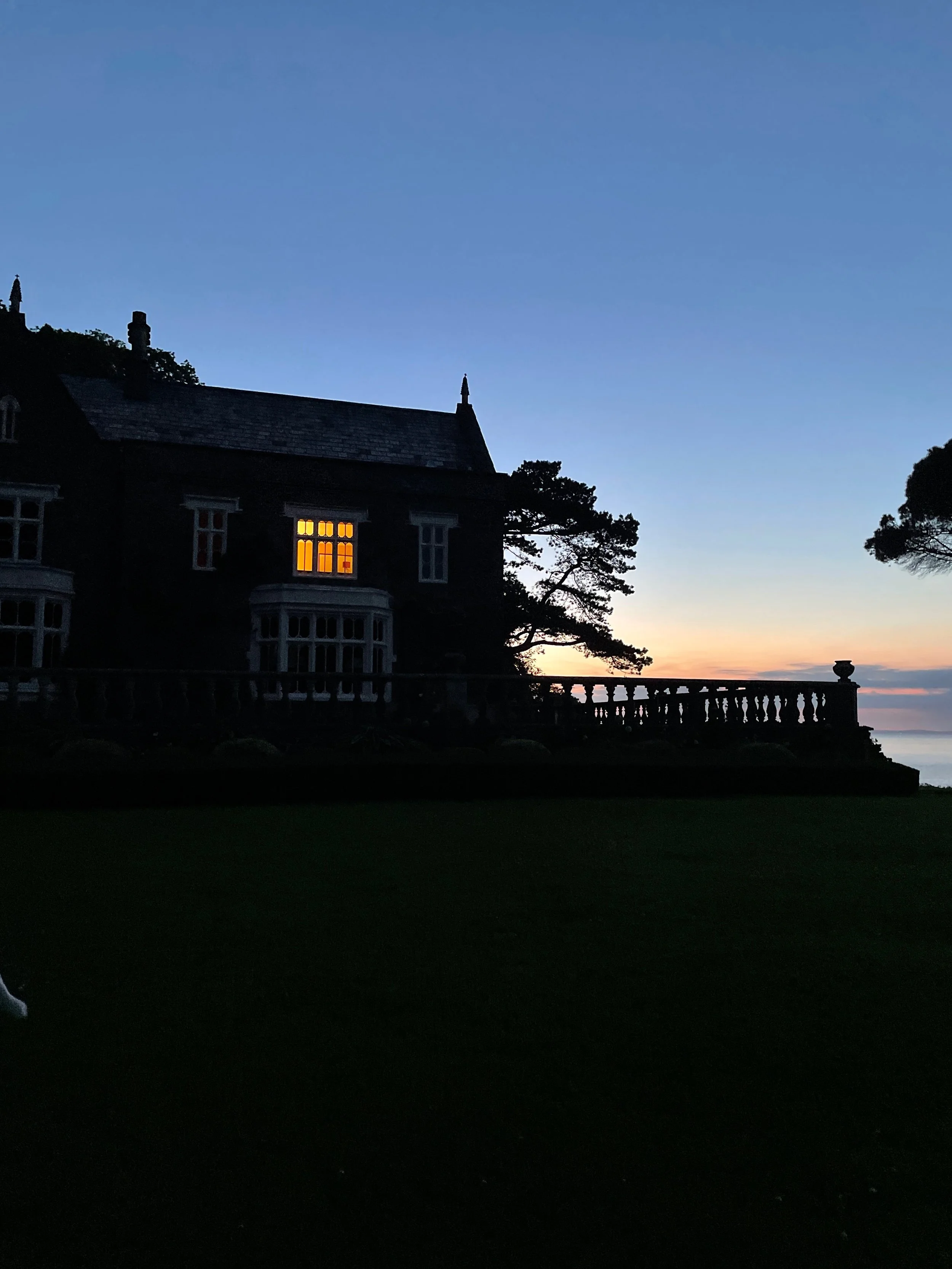A dark silhouette of a large house or mansion with a lit window, set against a colorful twilight sky with a hint of sunset on the horizon, trees, and a balustrade terrace.