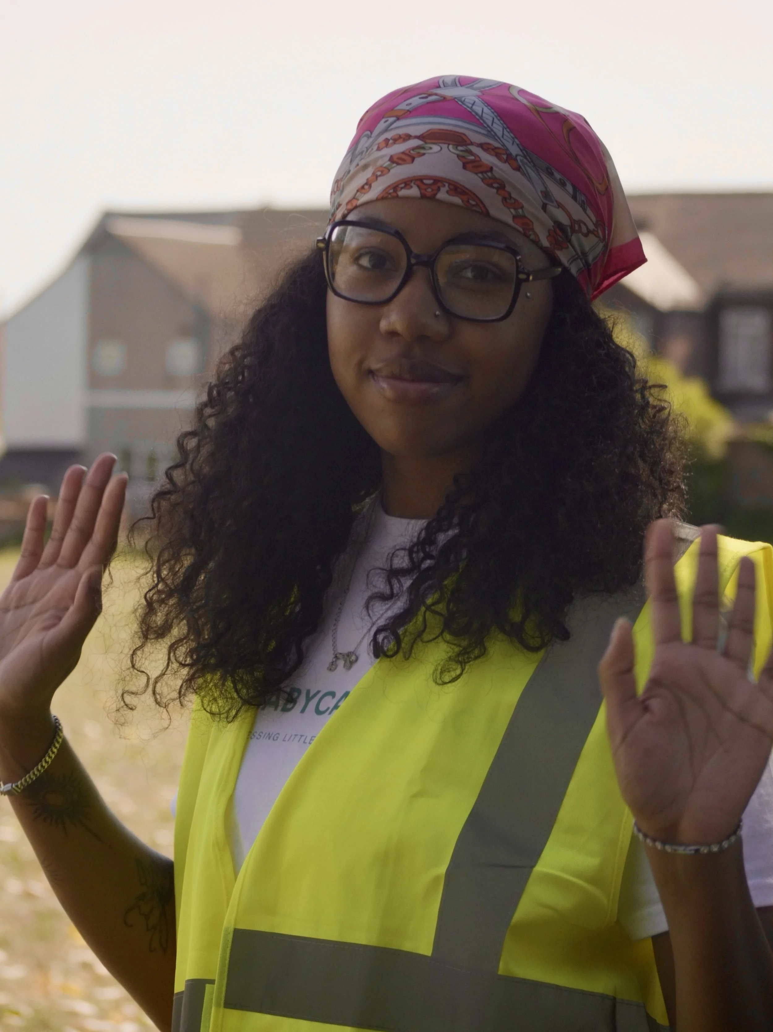 A woman wearing glasses and a pink bandana, raising her hands, standing outdoors in a residential neighborhood, wearing a yellow safety vest.