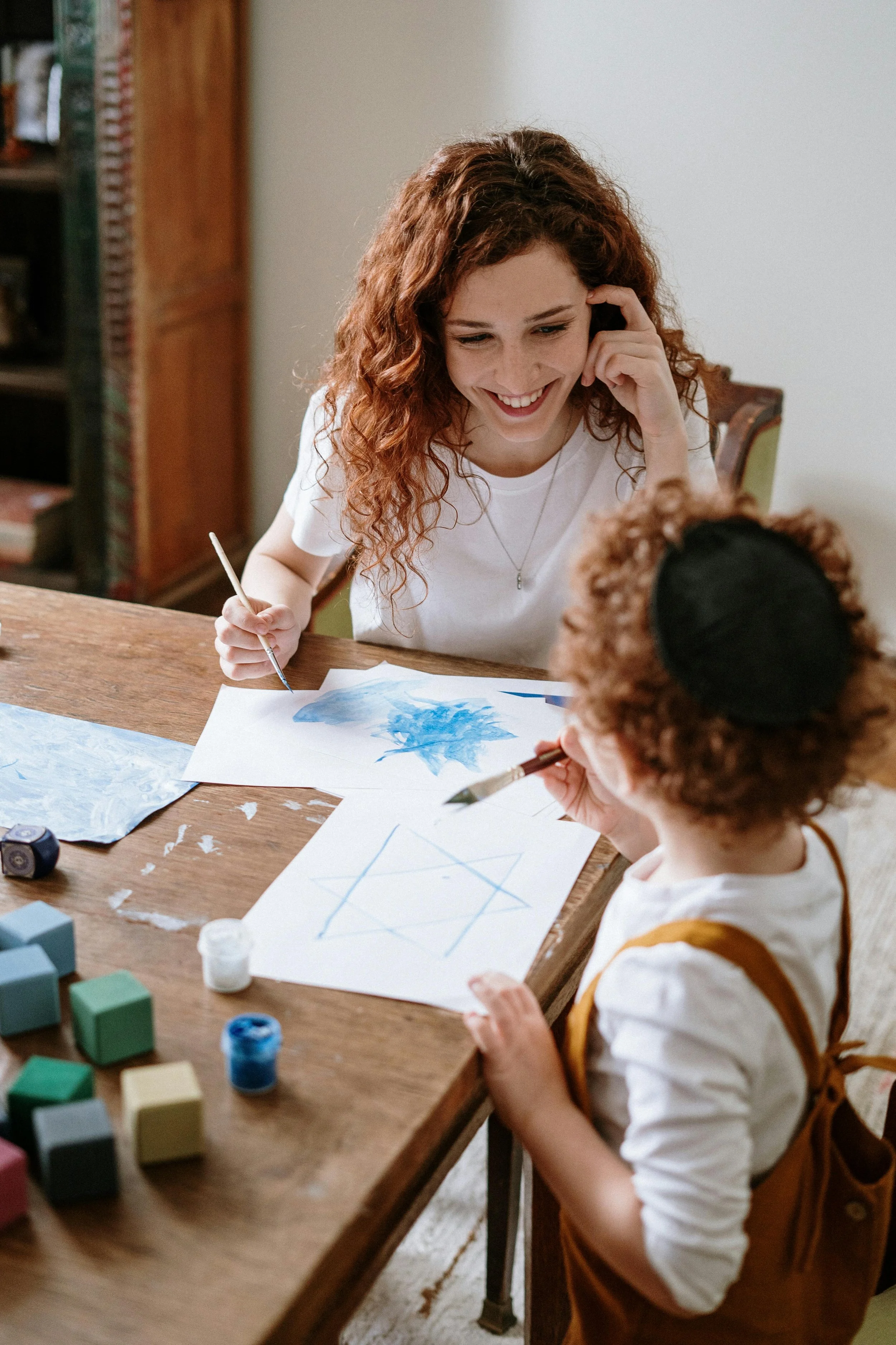 A woman and a young girl are sitting at a wooden table, painting and drawing together. The woman has curly red hair and is smiling, holding a paintbrush. The girl has curly hair with a hairband and is focused on her drawing. Art supplies, including paints and papers, are on the table.