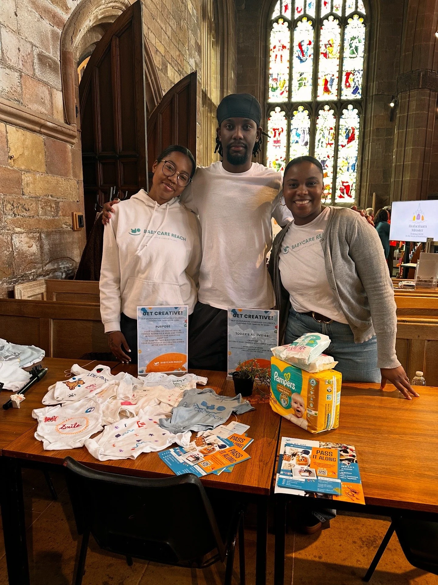Three people standing behind a table with baby clothes, flyers, and baby supplies, inside a church with stained glass windows in the background.