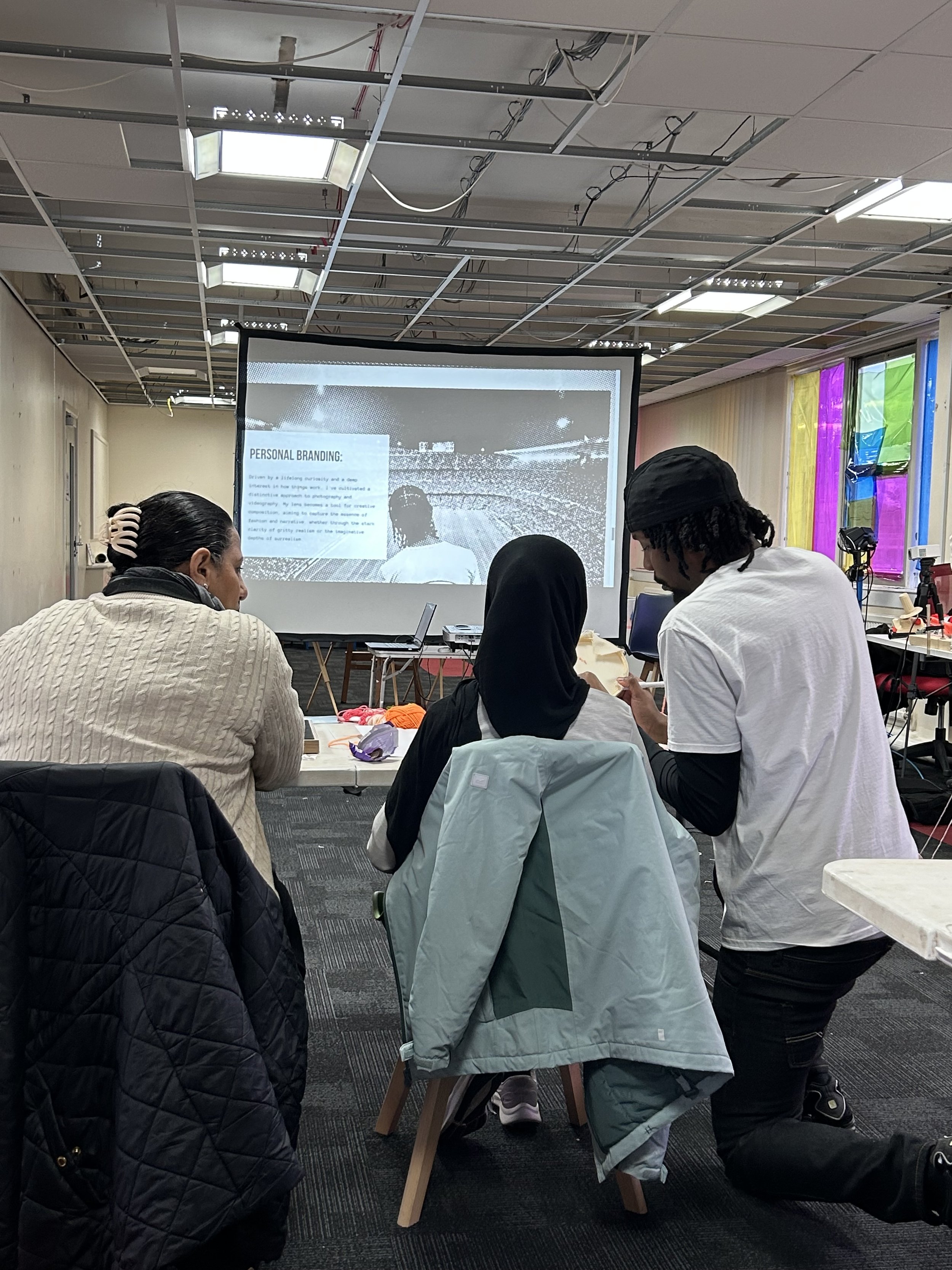 Three people sit at a table in front of a large screen displaying a presentation that reads "Personal Branding" with a stadium background. One person is standing and pointing at the screen, engaging with the other two attendees.