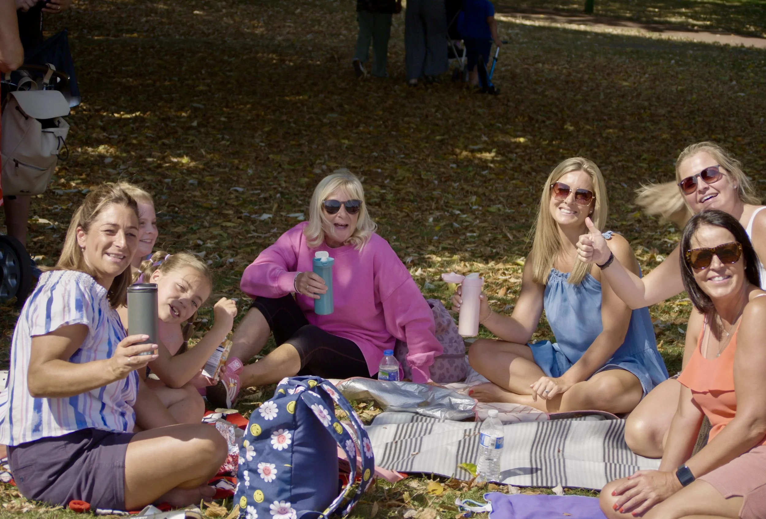 A group of women and a girl having a picnic outdoors on a blanket with drinks and snacks, surrounded by fallen leaves.