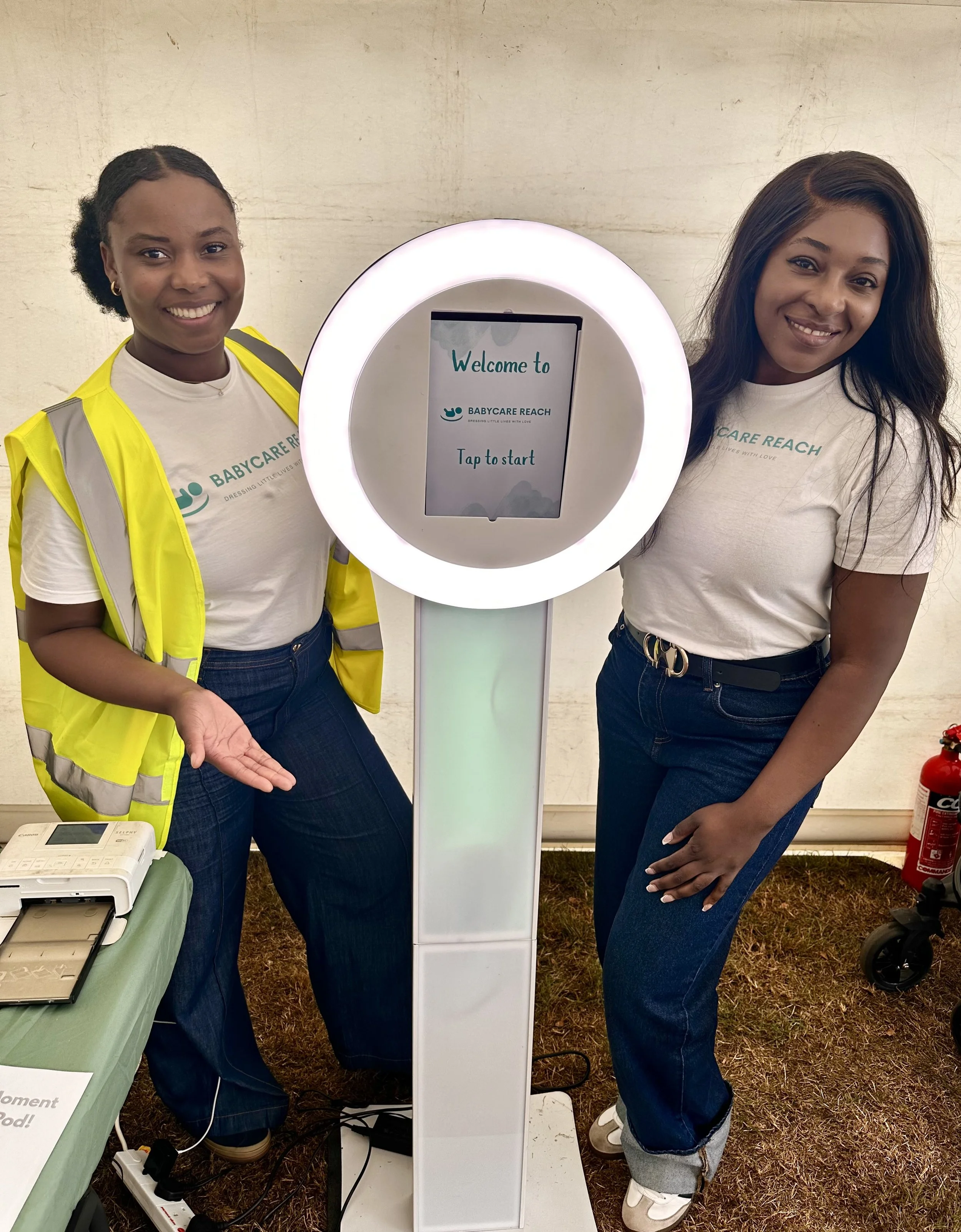 Two women standing beside a lit up kiosk with a circular opening, greeting to 'Welcome to BABYCARE REACH' on the screen, inside a tent.