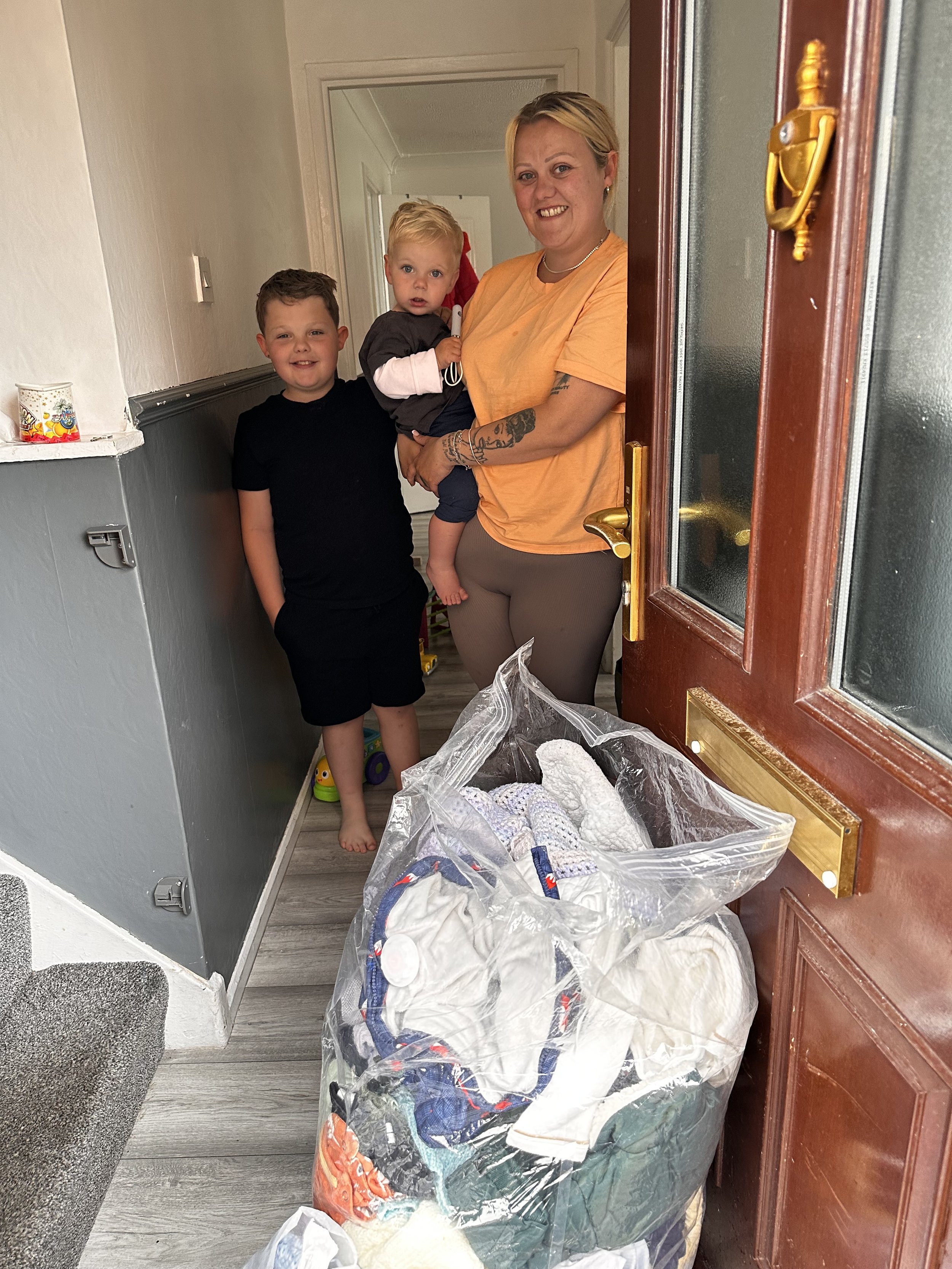 A woman with short blonde hair, wearing an orange shirt, stands at an open door with two young boys, one with short dark hair and the other with blonde hair, standing inside. The woman is holding a small child with blonde hair. There is a large bag of laundry in the foreground.