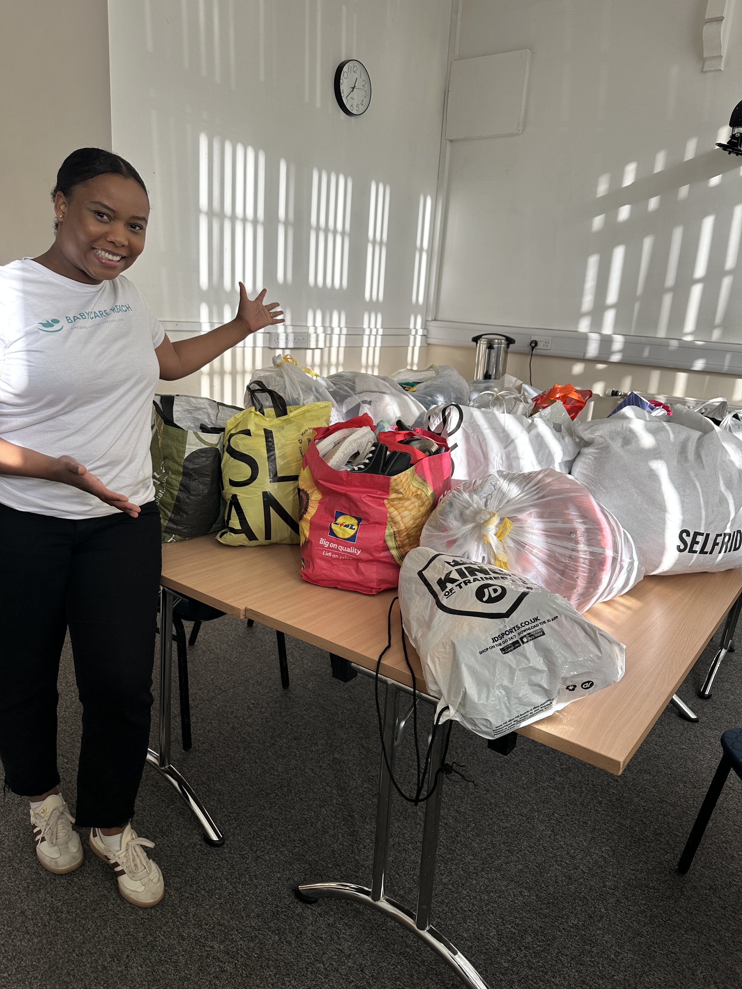 A woman standing next to a table filled with bags, some branded, in a room with sunlight and shadows from window blinds on the white wall.