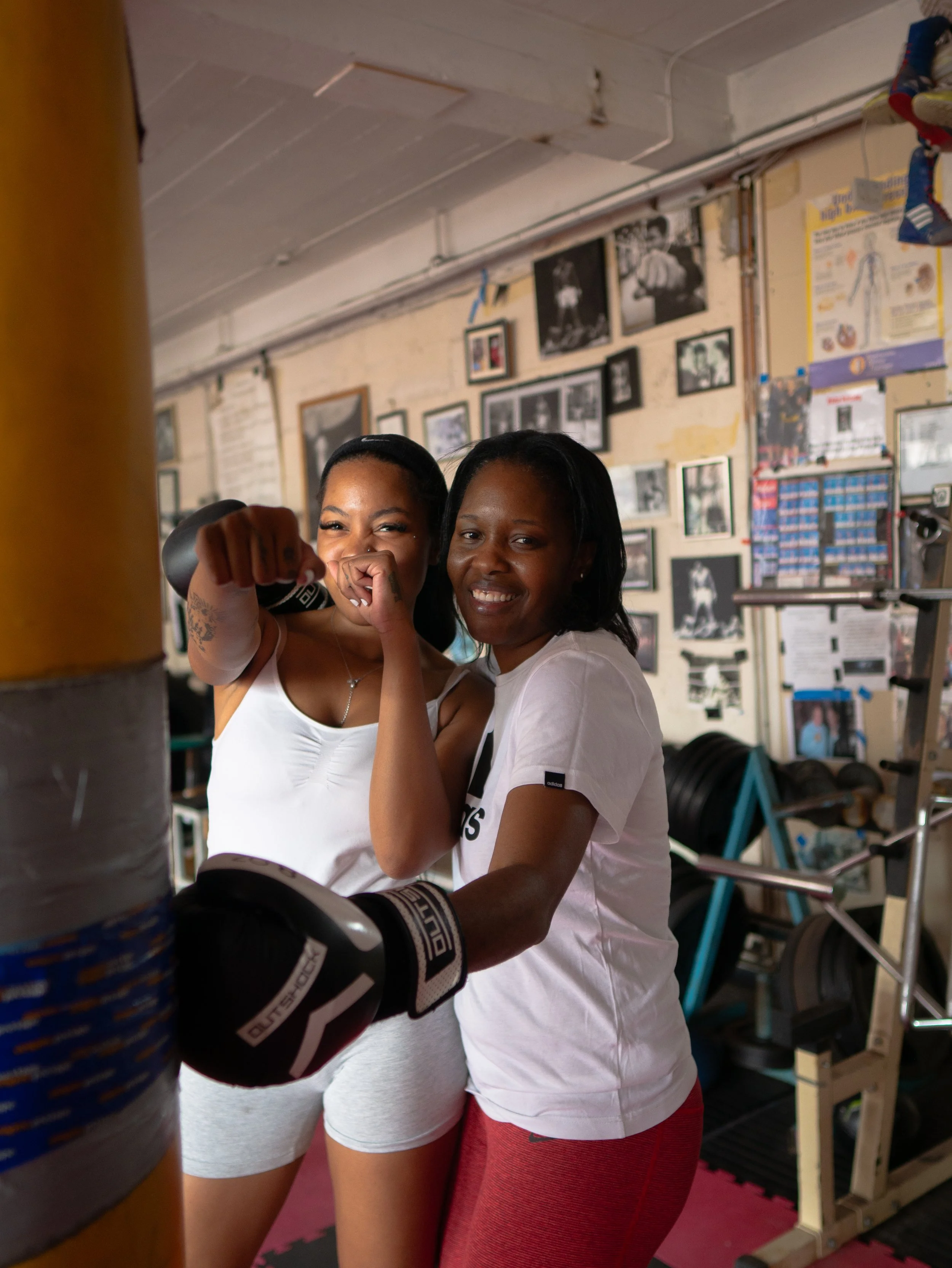 Two women in a gym, one wearing boxing gloves, smiling and posing for a photo.