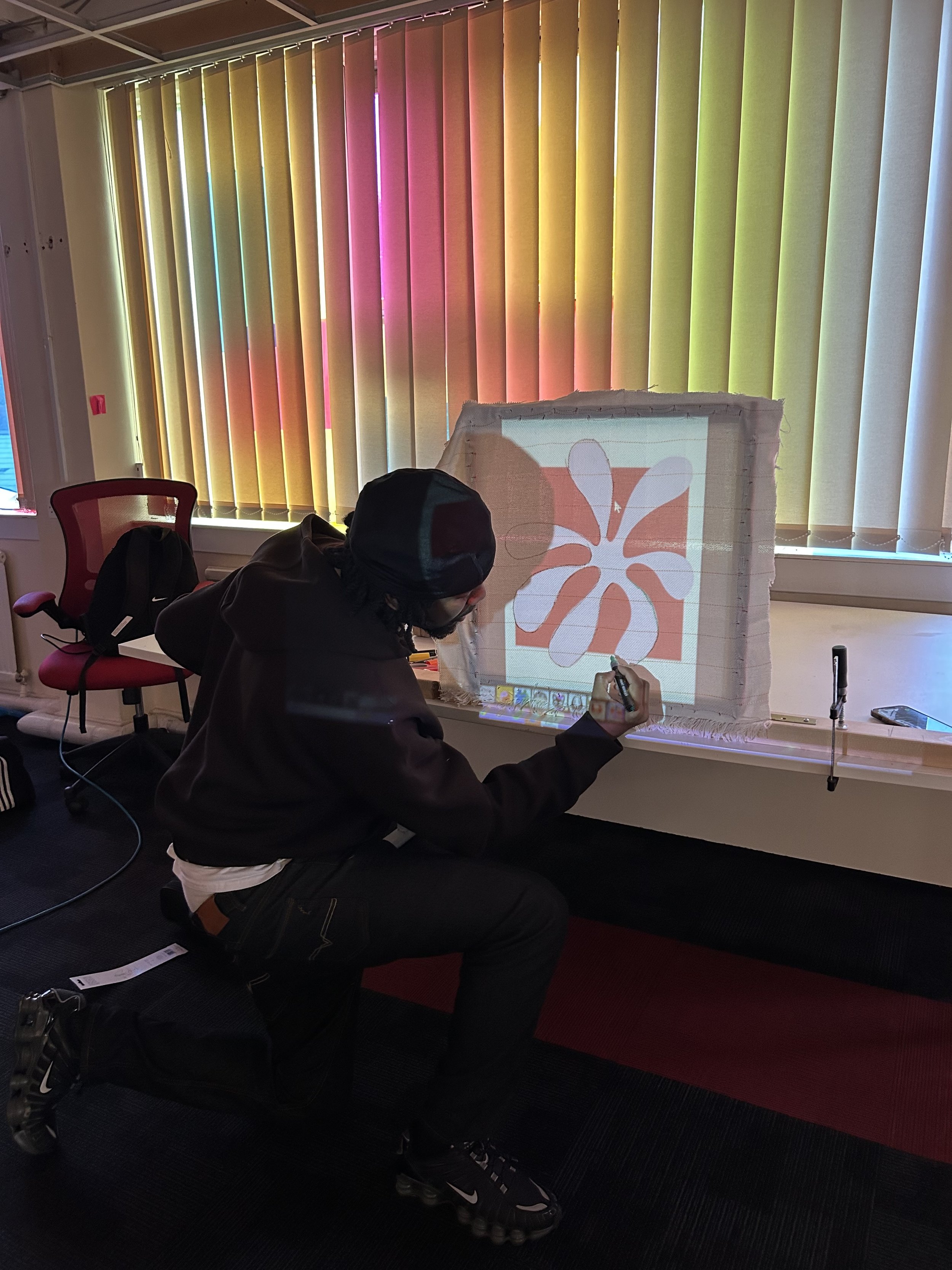 Person sitting on a black and red carpeted floor drawing on a whiteboard with a marker, in front of colorful vertical window blinds.