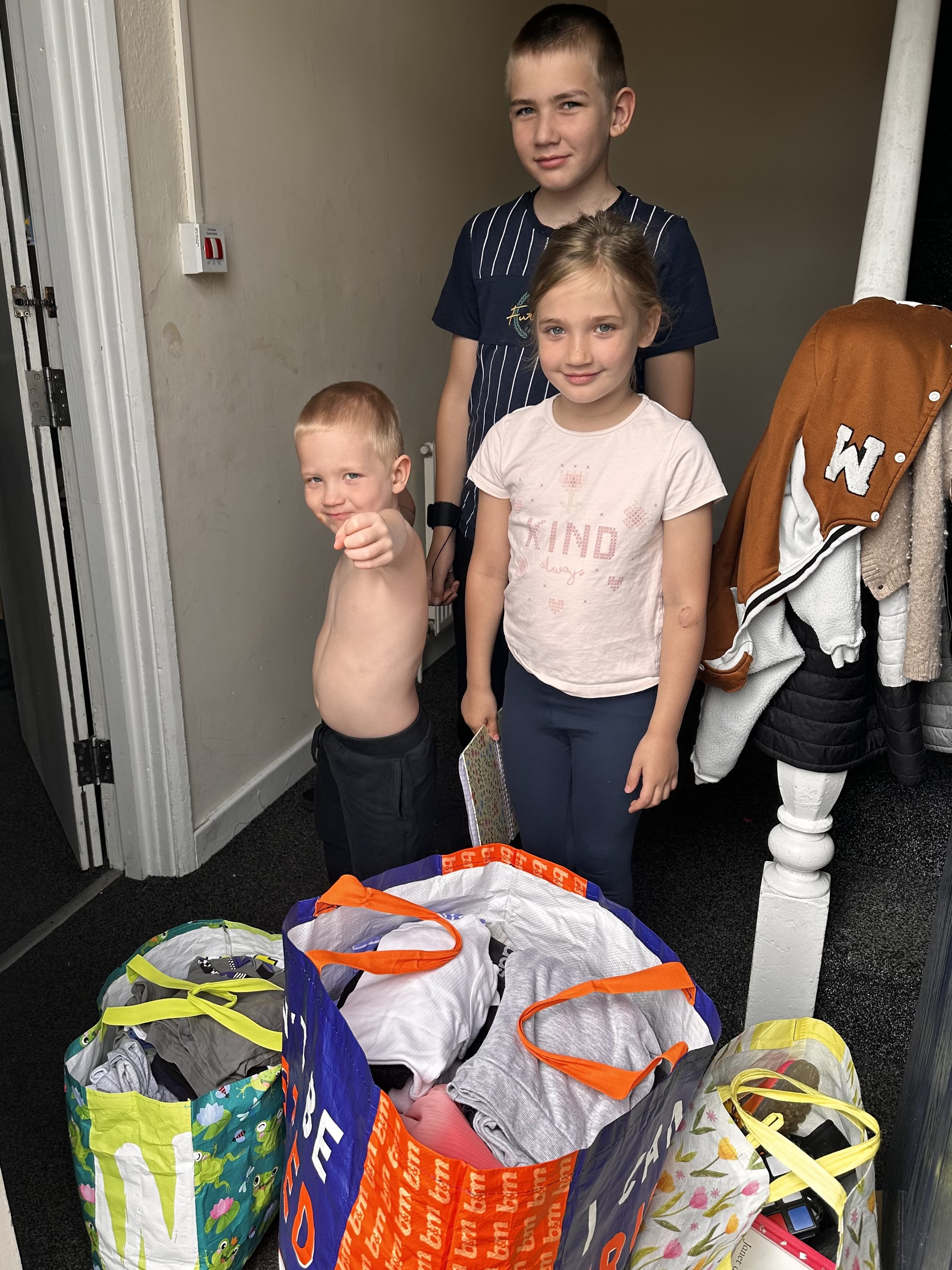 Four children standing inside a house, three girls and one boy, with shopping bags filled with clothes in front of them.