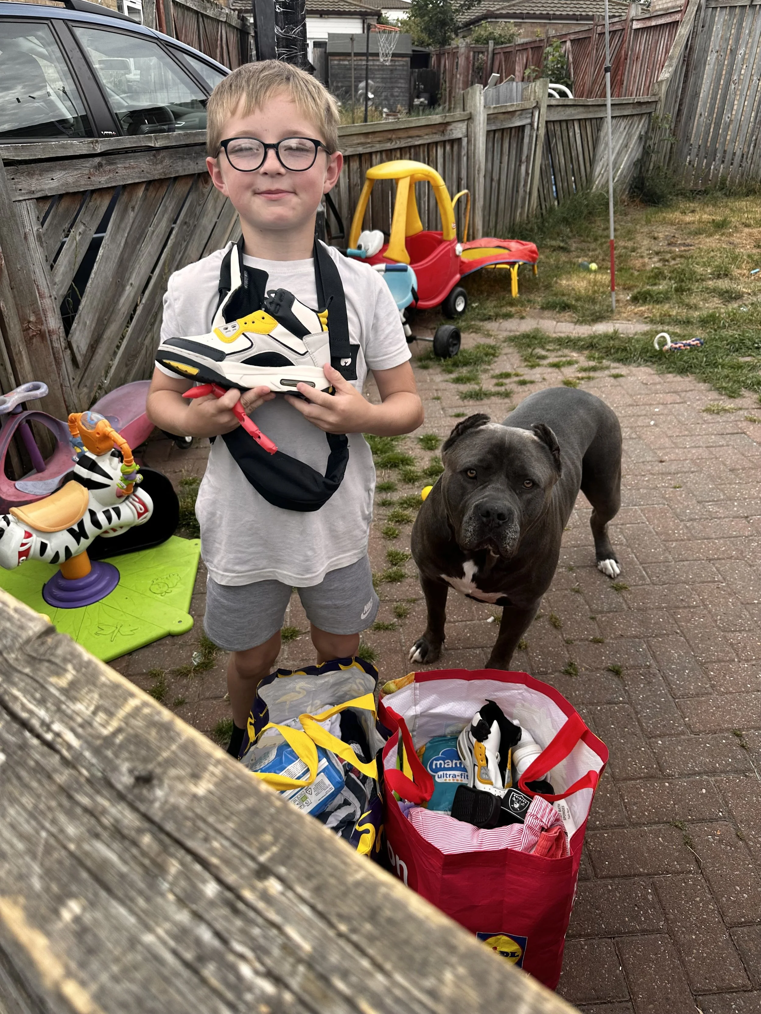 A young boy with glasses holding a pair of black, white, and yellow sneakers, standing outdoors in a backyard next to a gray pit bull dog. The yard has toys and bags on the ground, a wooden fence, a parked car, and a small red and yellow toy car in the background.