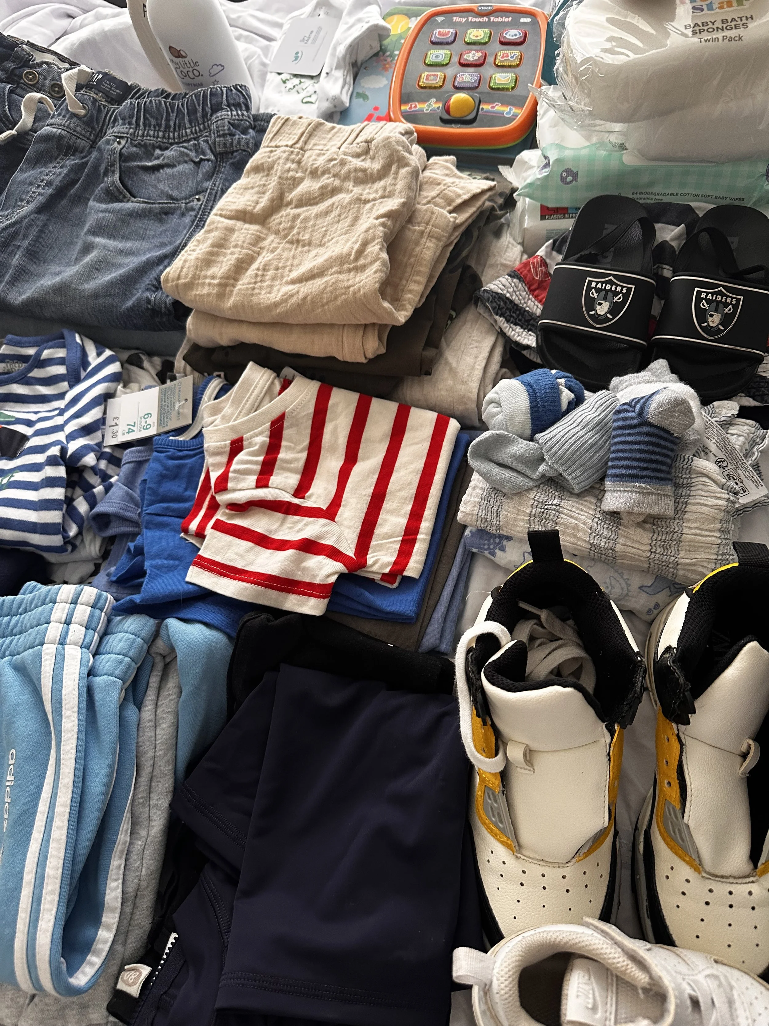 Collection of children's clothes, shoes, socks, and accessories, including jeans, shorts, striped shirts, and athletic shoes, along with toiletries, wipes, and toy tablets.