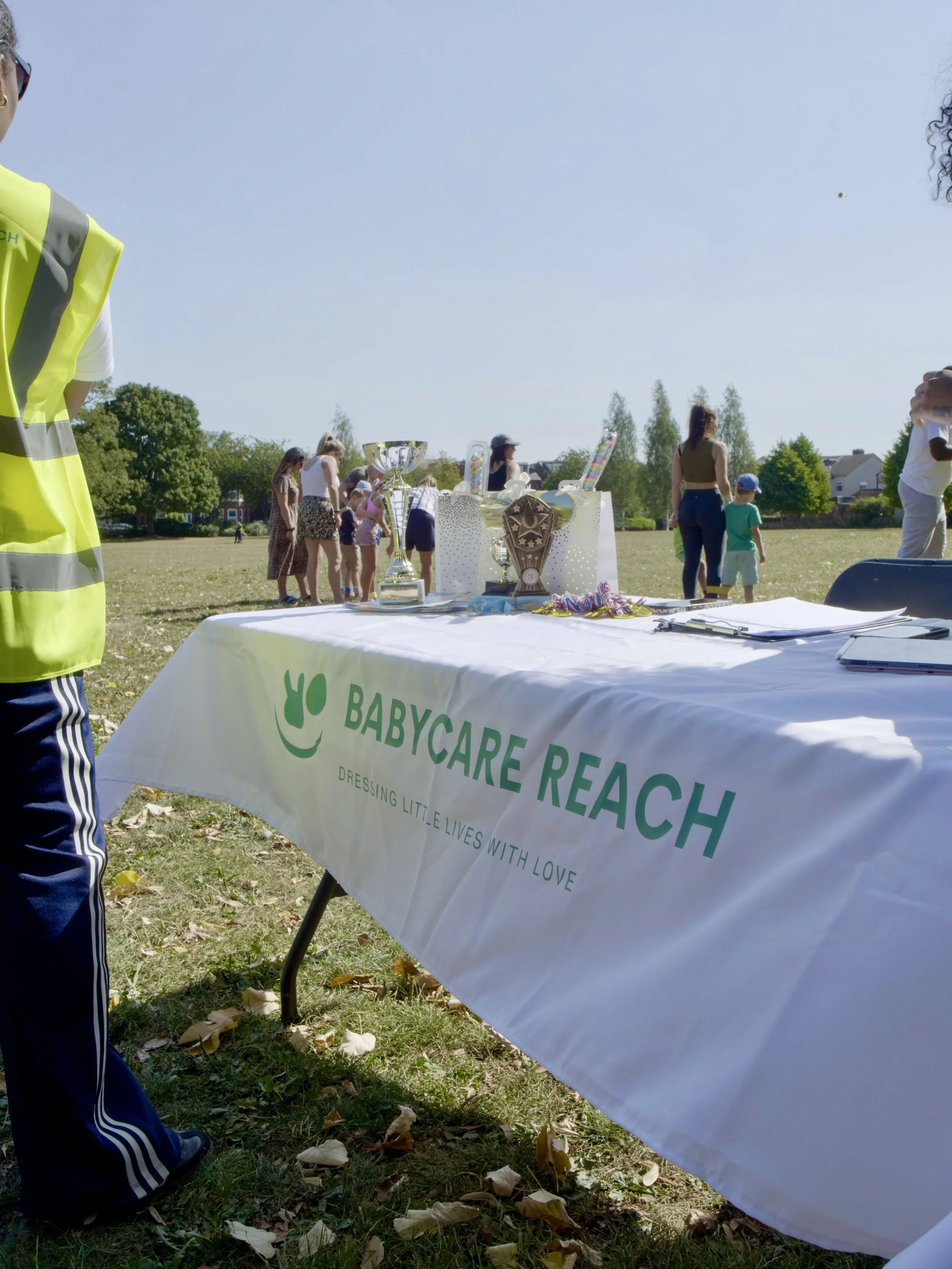 A table with a BabyCare Reach banner at an outdoor event, with trophies and awards on it, and children and adults in the background in a grassy park.