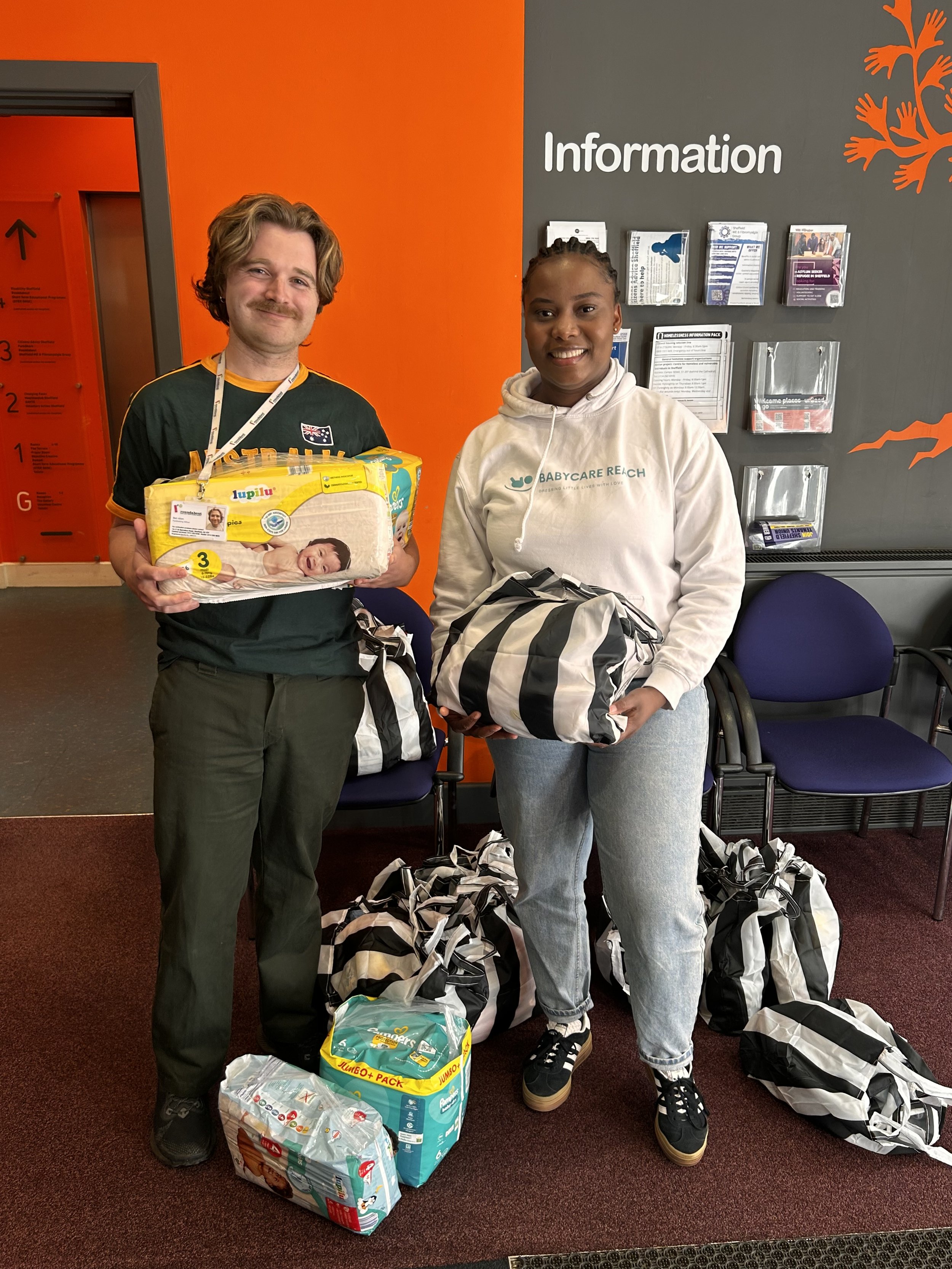Two people, a man and a woman, are standing in an indoor waiting area, holding packages and bags, with several more bags on the floor. They are smiling and appear to be at a healthcare or childcare facility, as indicated by a sign that says 'Information' and a logo on the woman's hoodie that reads 'Babycare Reach.' The man is holding a package of diapers and wearing a dark shirt with a lanyard, while the woman is dressed casually in a white hoodie and jeans.