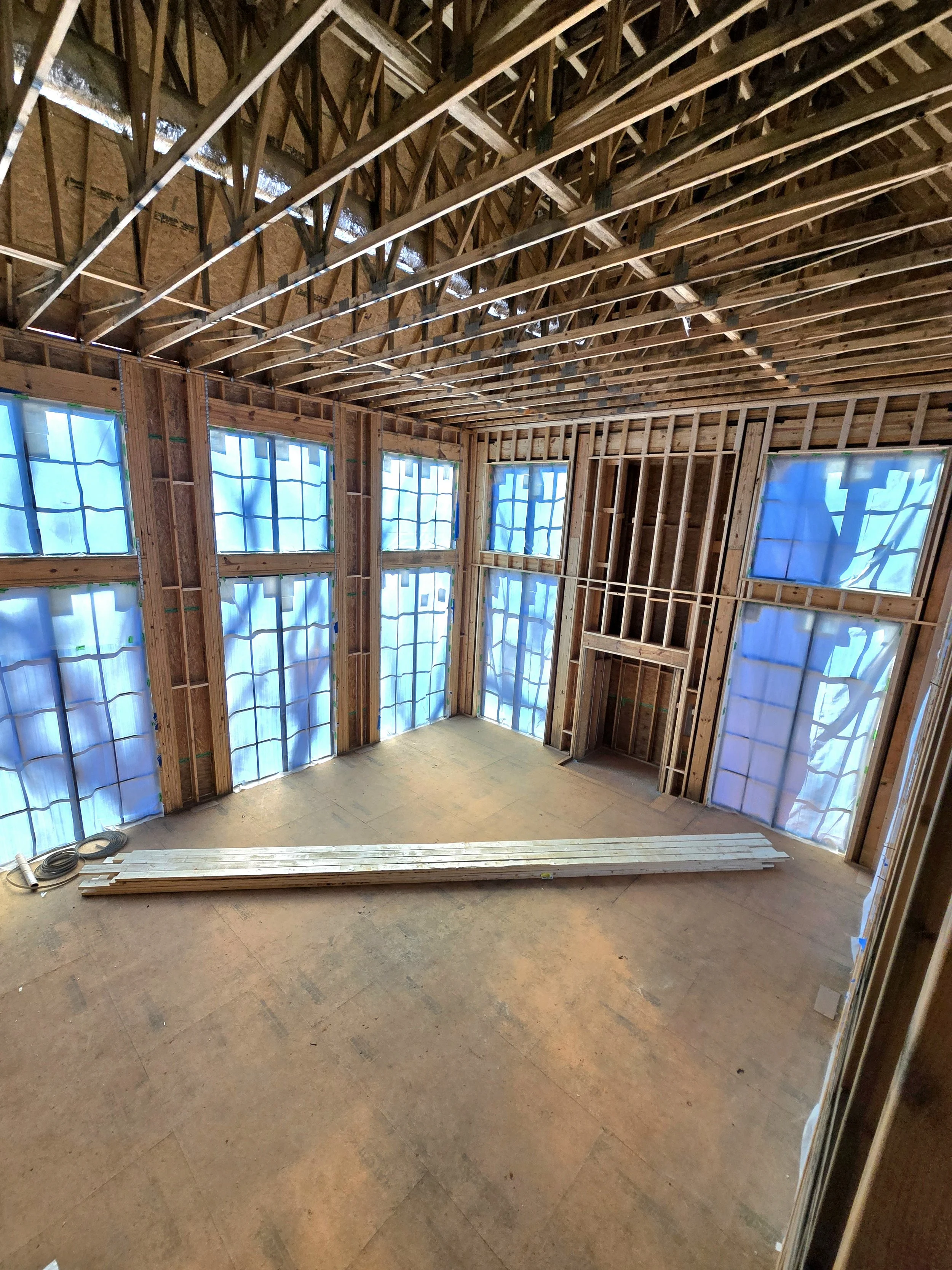 Interior of a house under construction with exposed wooden framing, partially covered windows with blue plastic sheeting, and a plywood floor, with some lumber stacked on the floor.