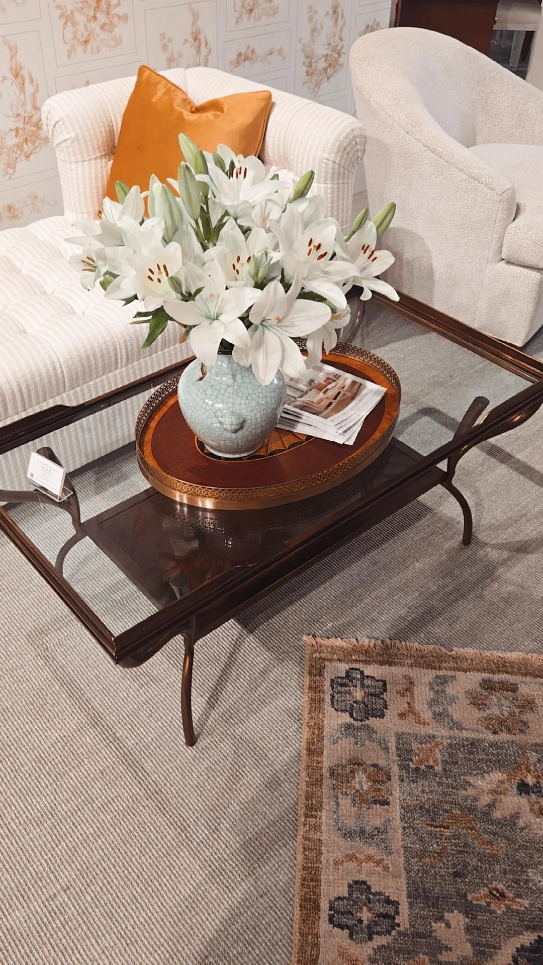 A living room scene with a glass coffee table holding a large white lily flower arrangement in a blue and crackled vase. The table is on a beige and gray area rug, with a cream-colored sofa and a beige armchair in the background. An orange pillow is on the sofa, and a magazine is next to the flowers.