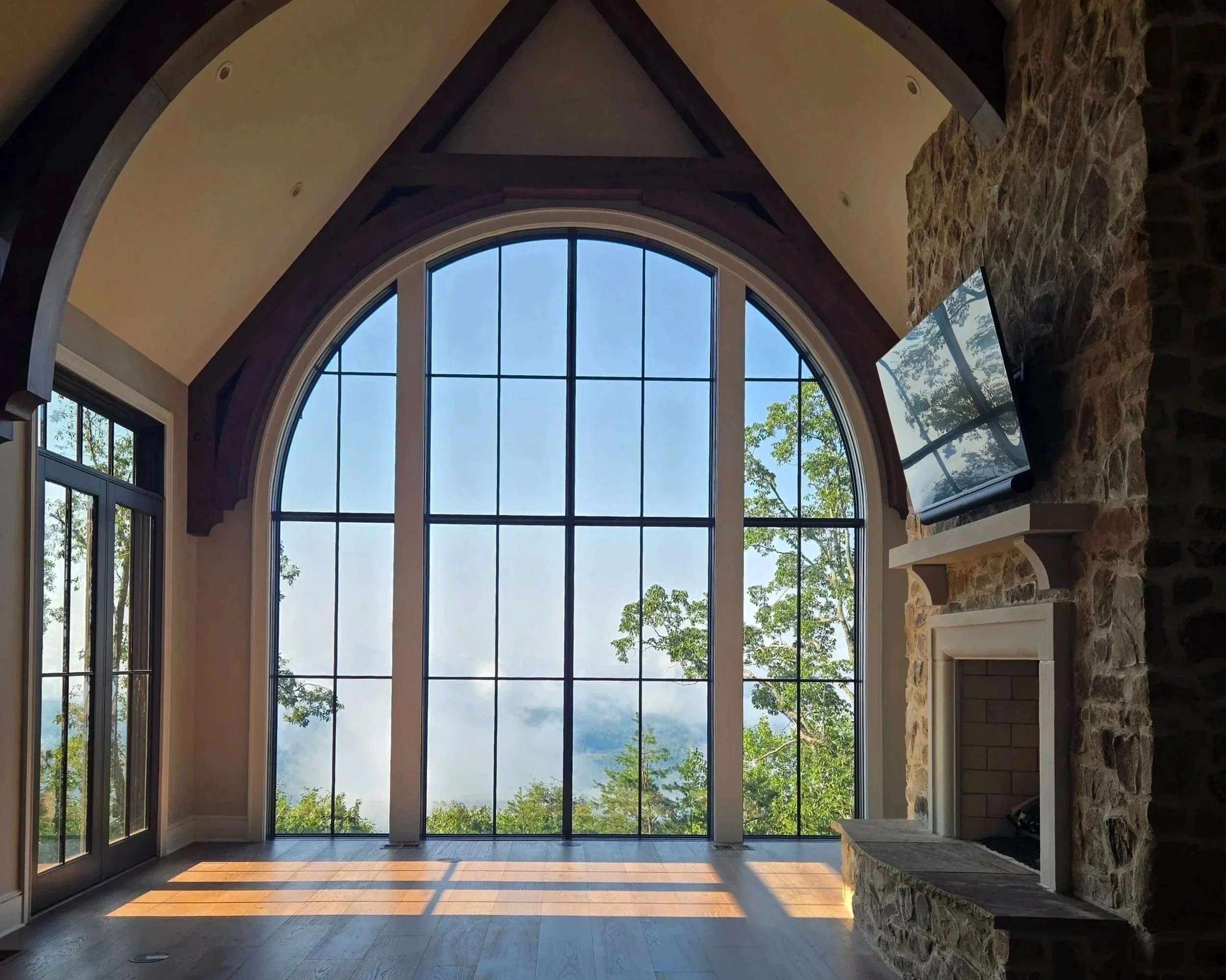 Large arched window with mountain and tree view, stone fireplace, and mounted flat-screen TV in living room.