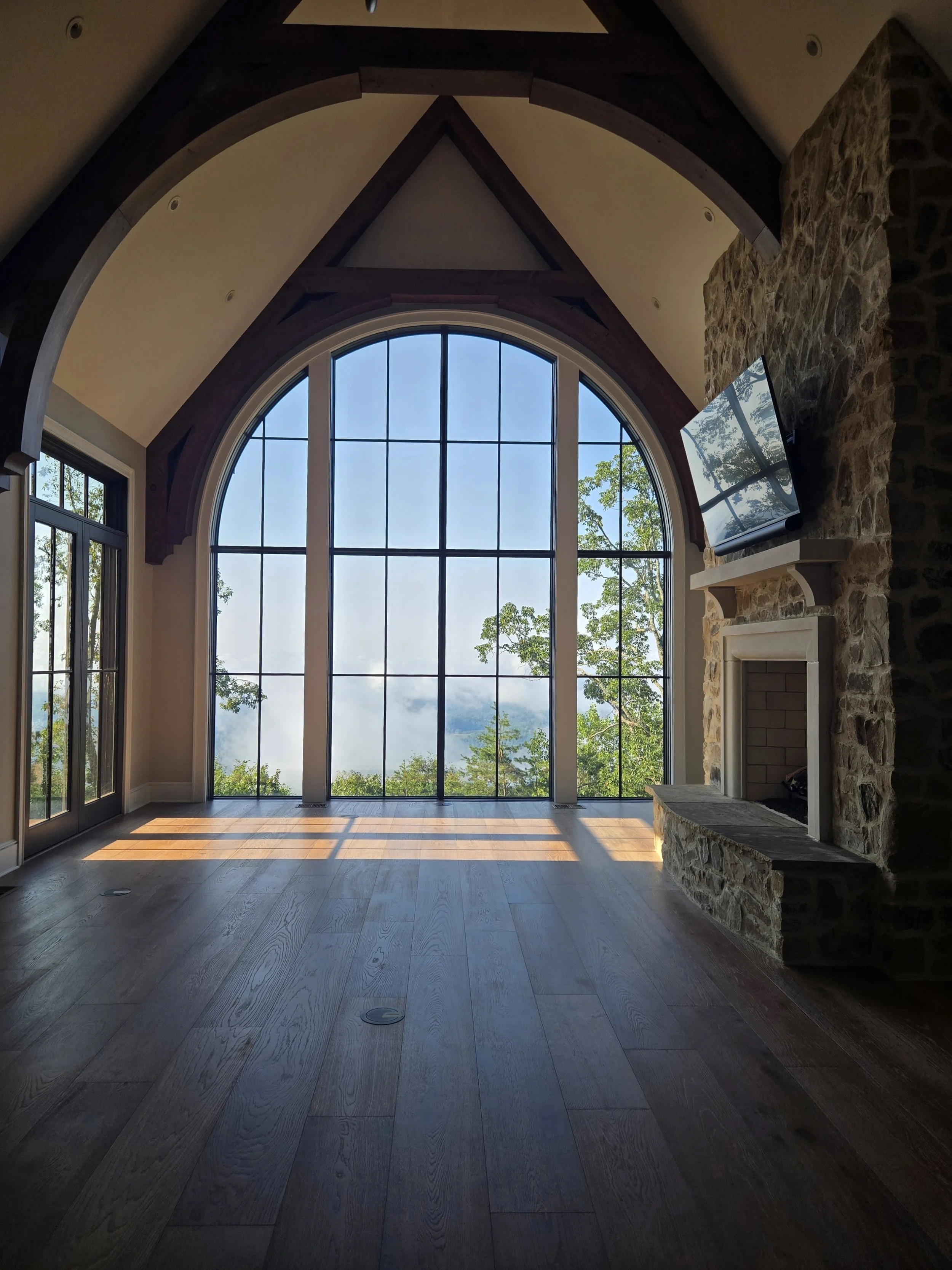 A spacious room with large arched window showing a view of trees and mountains, wooden floor, stone fireplace, and a wall-mounted flat-screen TV.