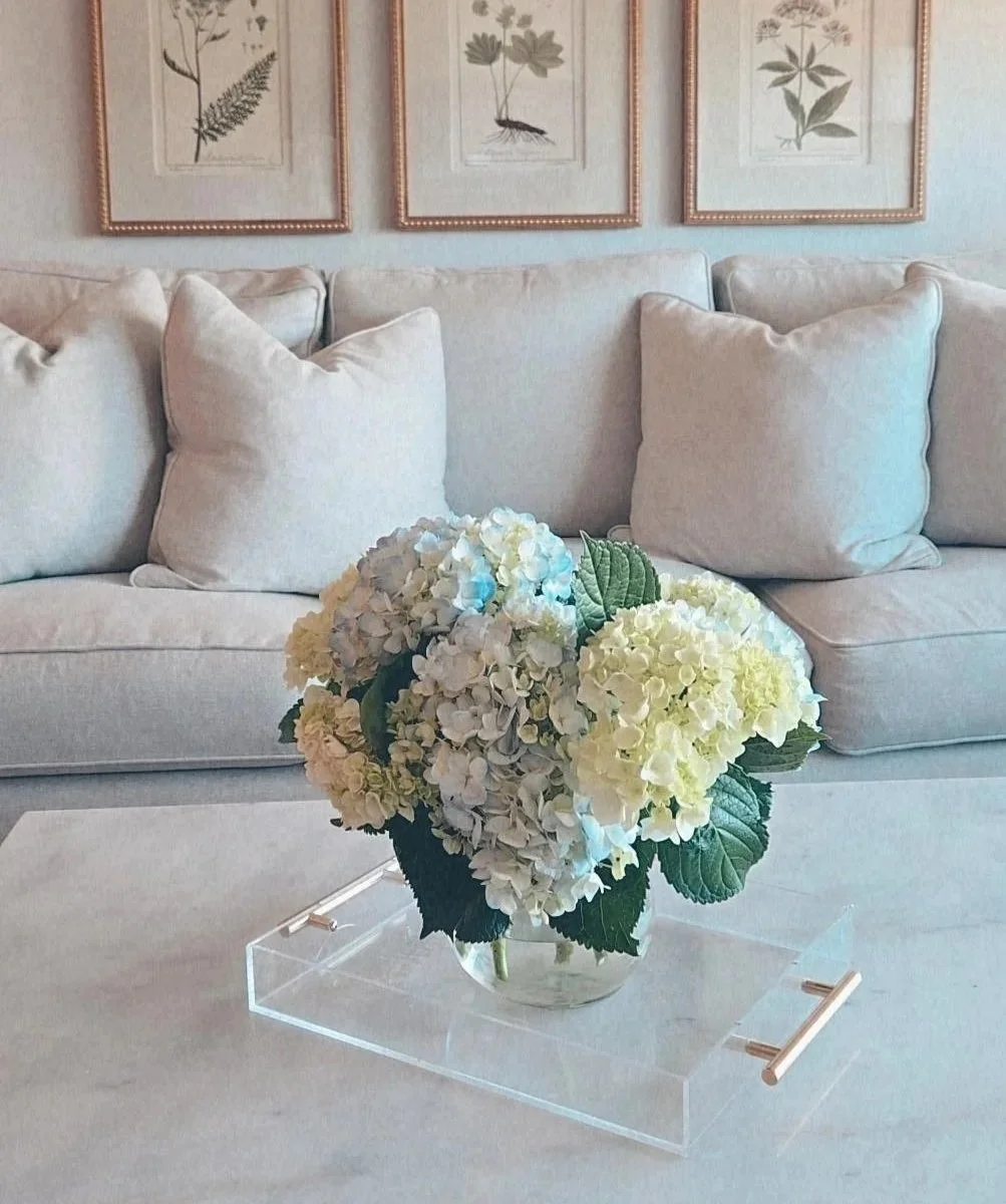 A beige sofa with multiple beige pillows, a glass coffee table with a white hydrangea flower arrangement in a clear vase, and botanical prints hanging on the wall.