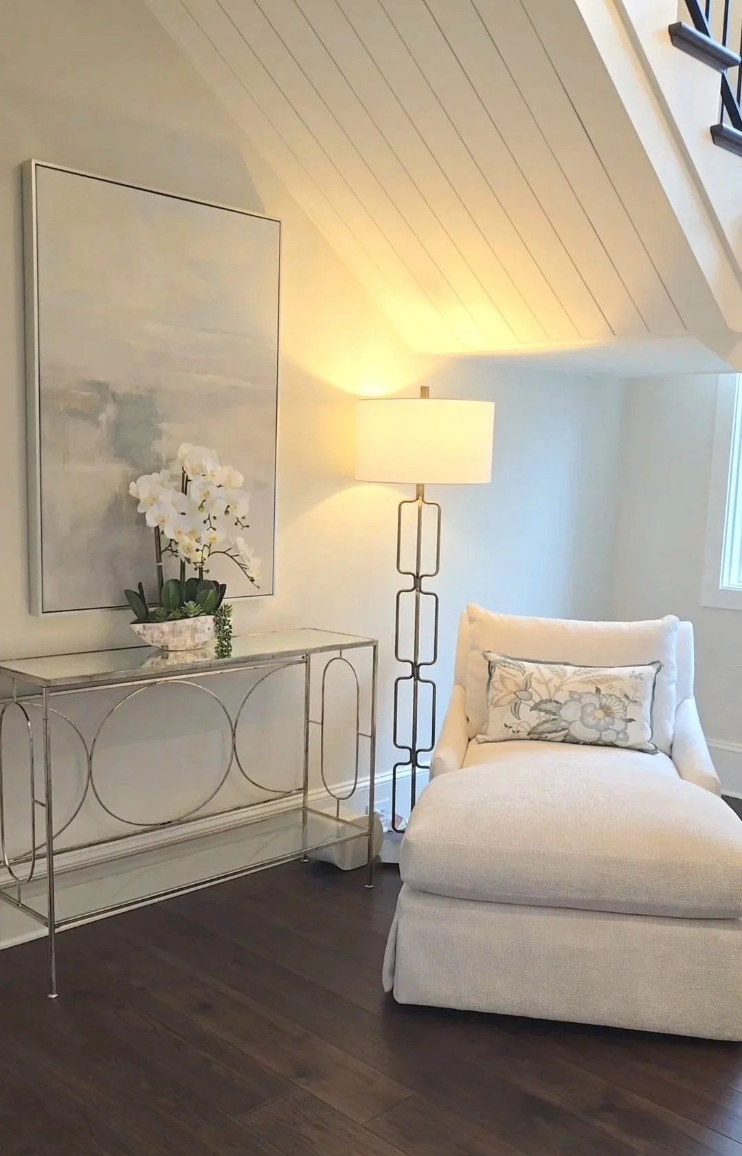 A living room corner with a white chaise lounge with a floral pillow, a tall floor lamp with a white shade, a glass and metal console table with a potted orchid, a large abstract painting, and a window with sunlight.