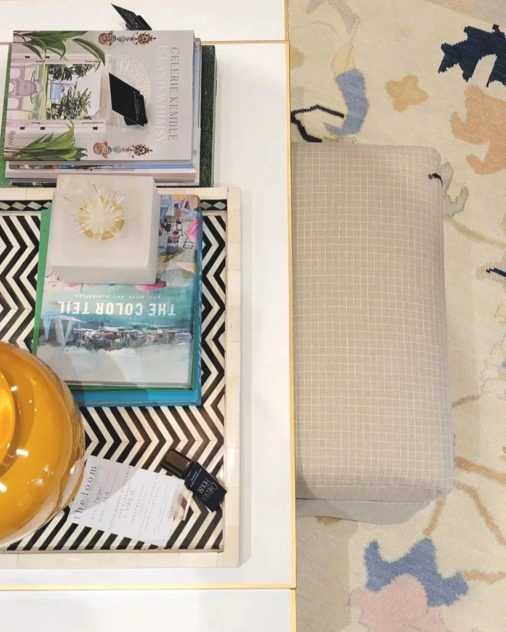 A table with decorative items, fashion magazines, a tissue box, a large yellow vase, and a receipt, with a beige checkered ottoman and a colorful patterned rug underneath.