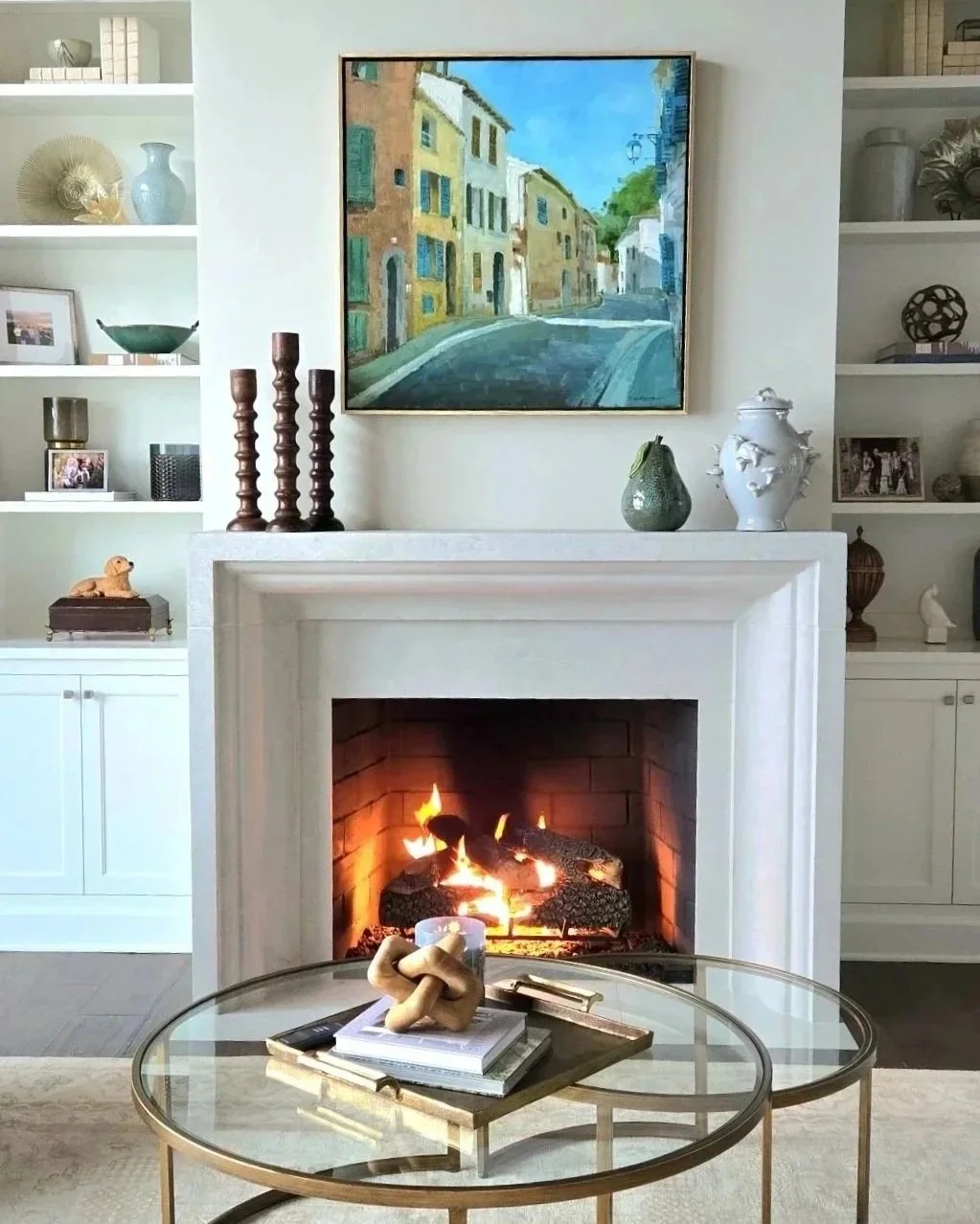 Living room with a white fireplace, lit fire inside, artwork of a street scene hanging above the fireplace, and decorative items on the mantel and shelves.