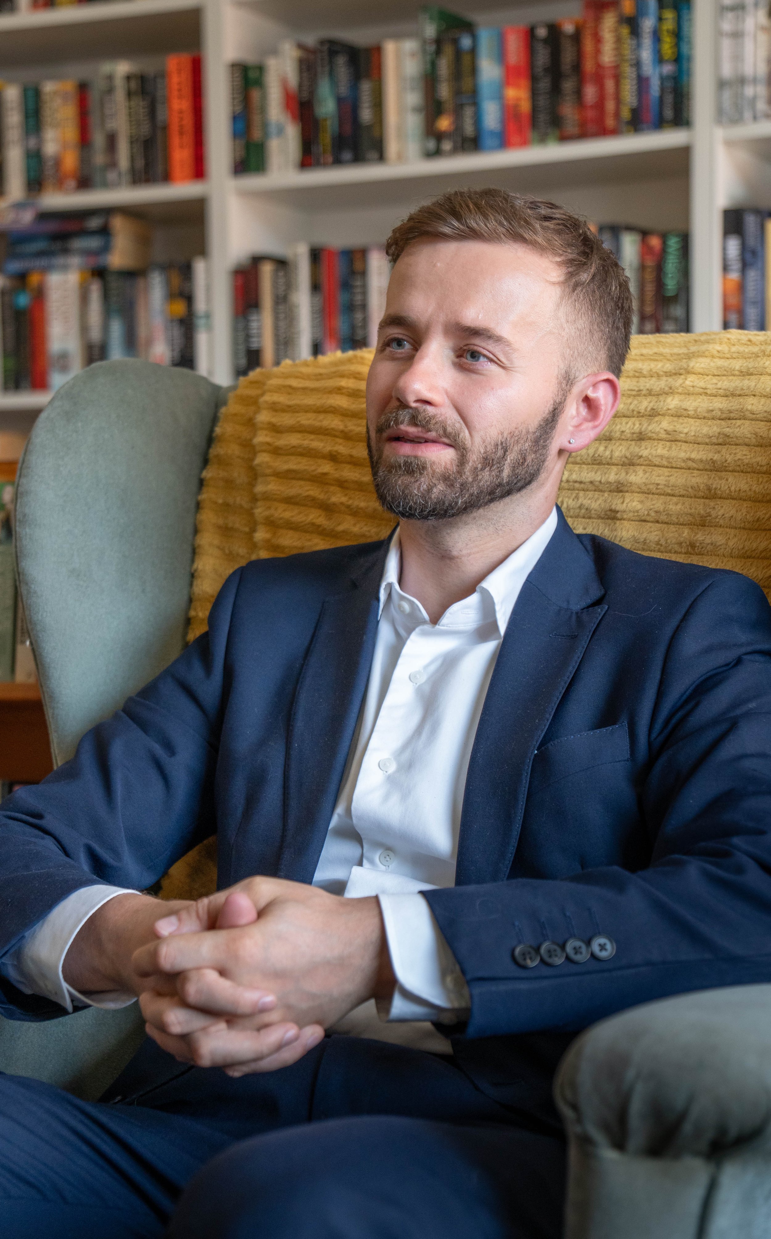 Portrait of English tutor Timothy Lunn sitting with hands clasped, bookshelves behind him