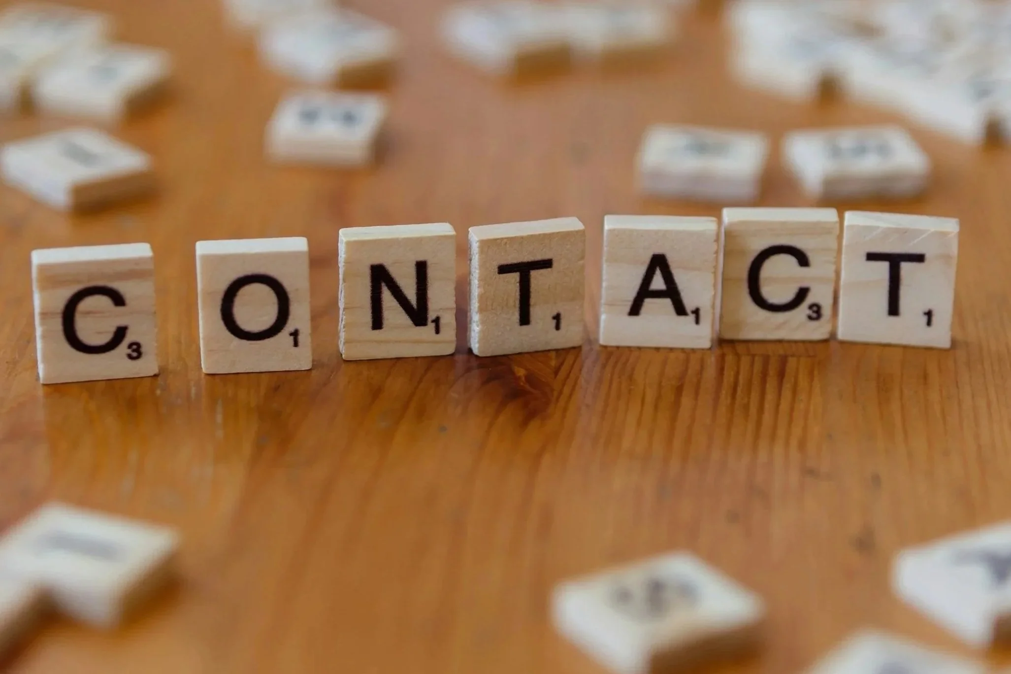 Scrabble tiles spelling out the word 'CONTACT' on a wooden table surrounded by scattered tiles.