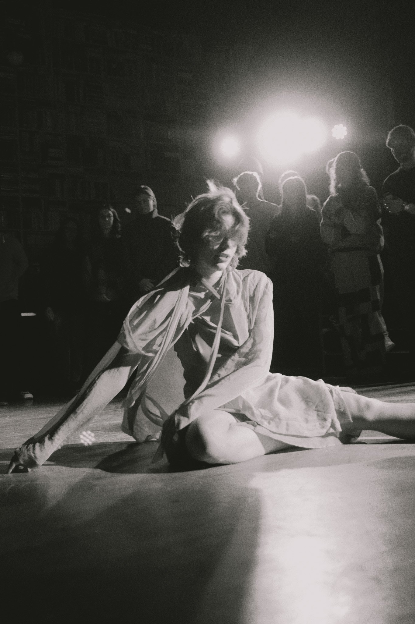 A woman in a dress is sitting on the floor with one hand touching the ground and the other resting on her knee, surrounded by a group of people in the background in a dimly lit space with bright lights overhead.