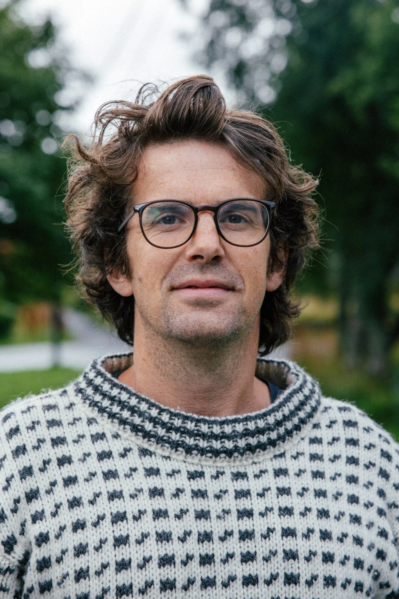 A man with curly brown hair, wearing glasses and a black and white patterned sweater, standing outdoors with trees and a body of water in the background.