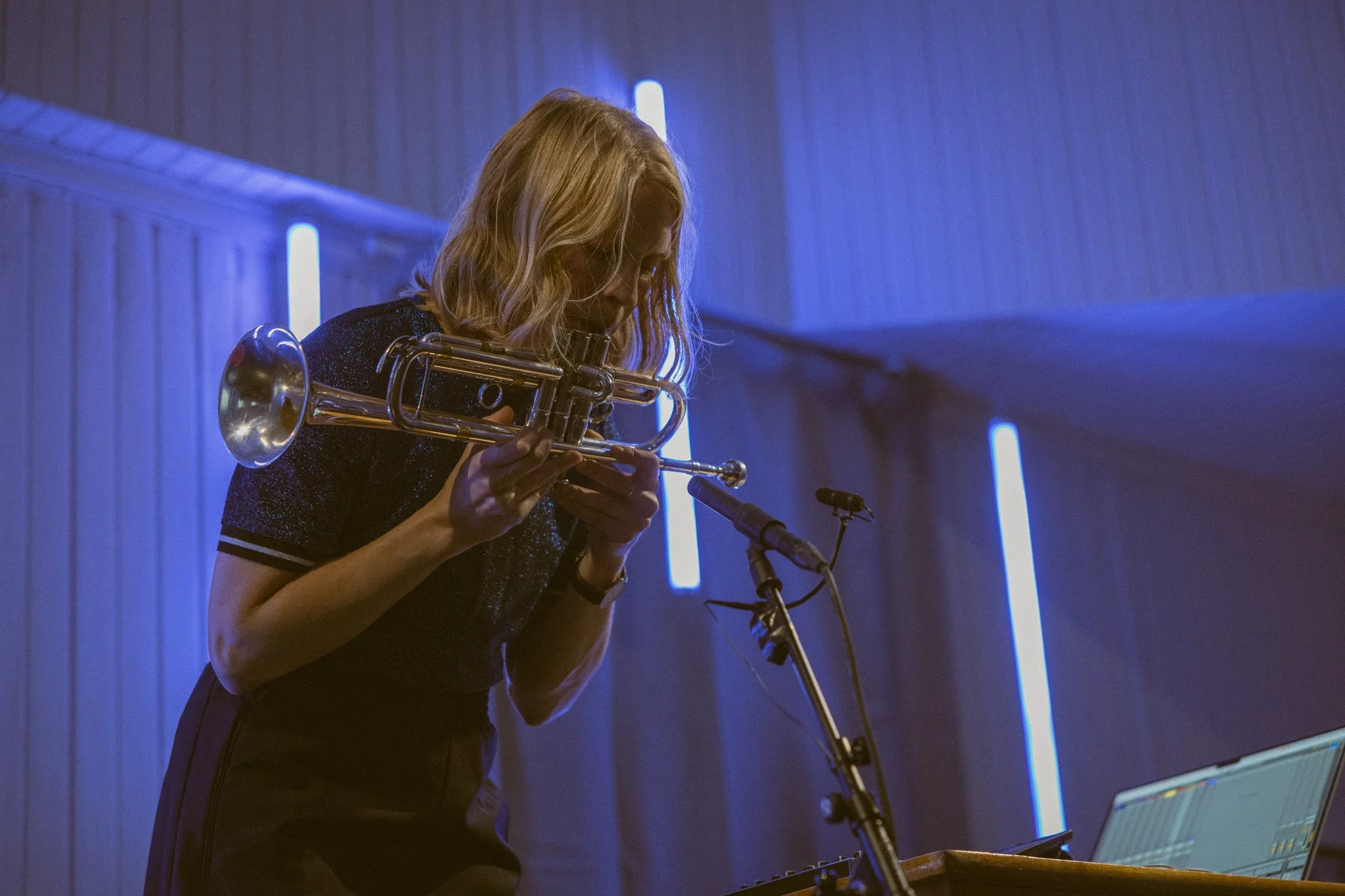 A woman with blonde hair, wearing a dark top, playing a trumpet in a dimly lit room with blue lighting, using a microphone and a laptop on a stand.