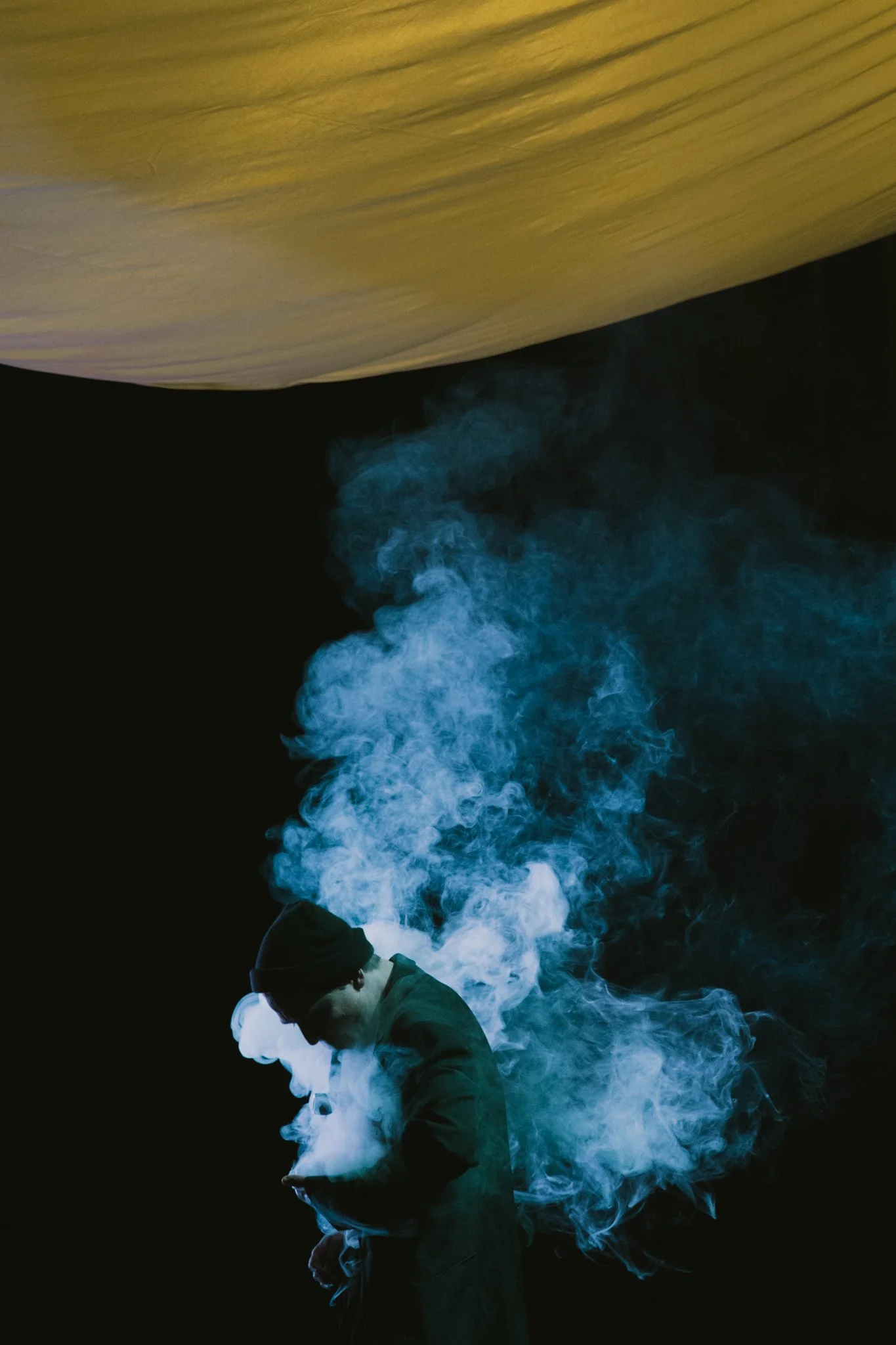 Person wearing a black beanie and coat, surrounded by blue and white smoke, against a dark background.