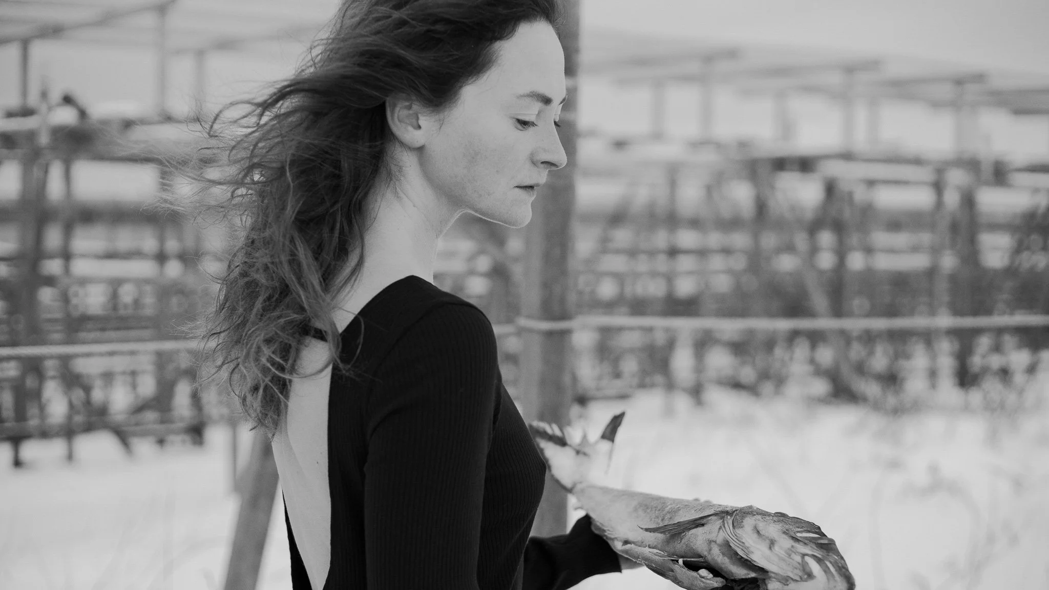 A woman with wavy hair in a black long-sleeve dress holding a bird, standing outdoors near some metal structures.