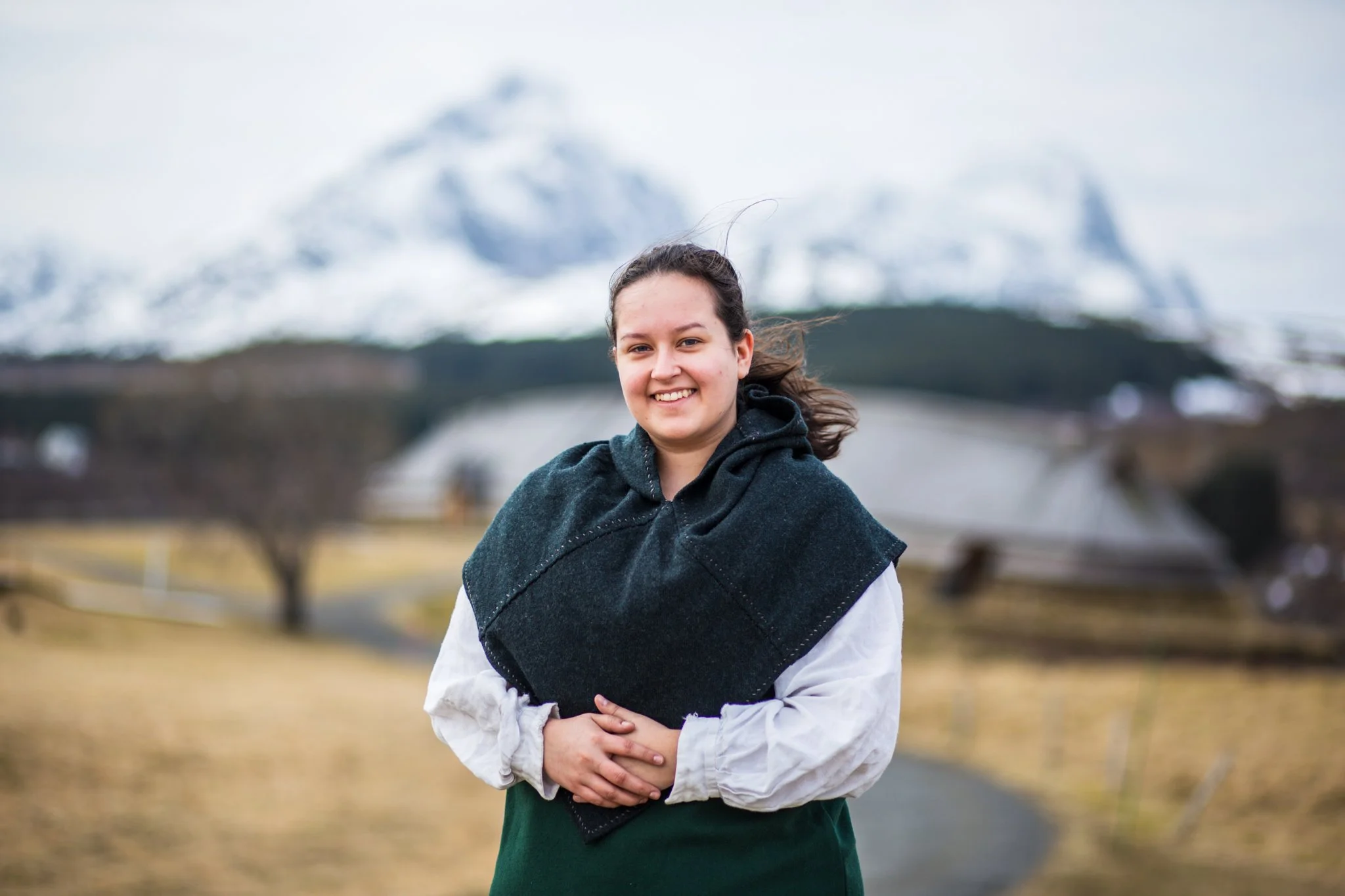 Young woman outdoors in front of snow-capped mountains, wearing a dark shawl and a white shirt, smiling with hands clasped.