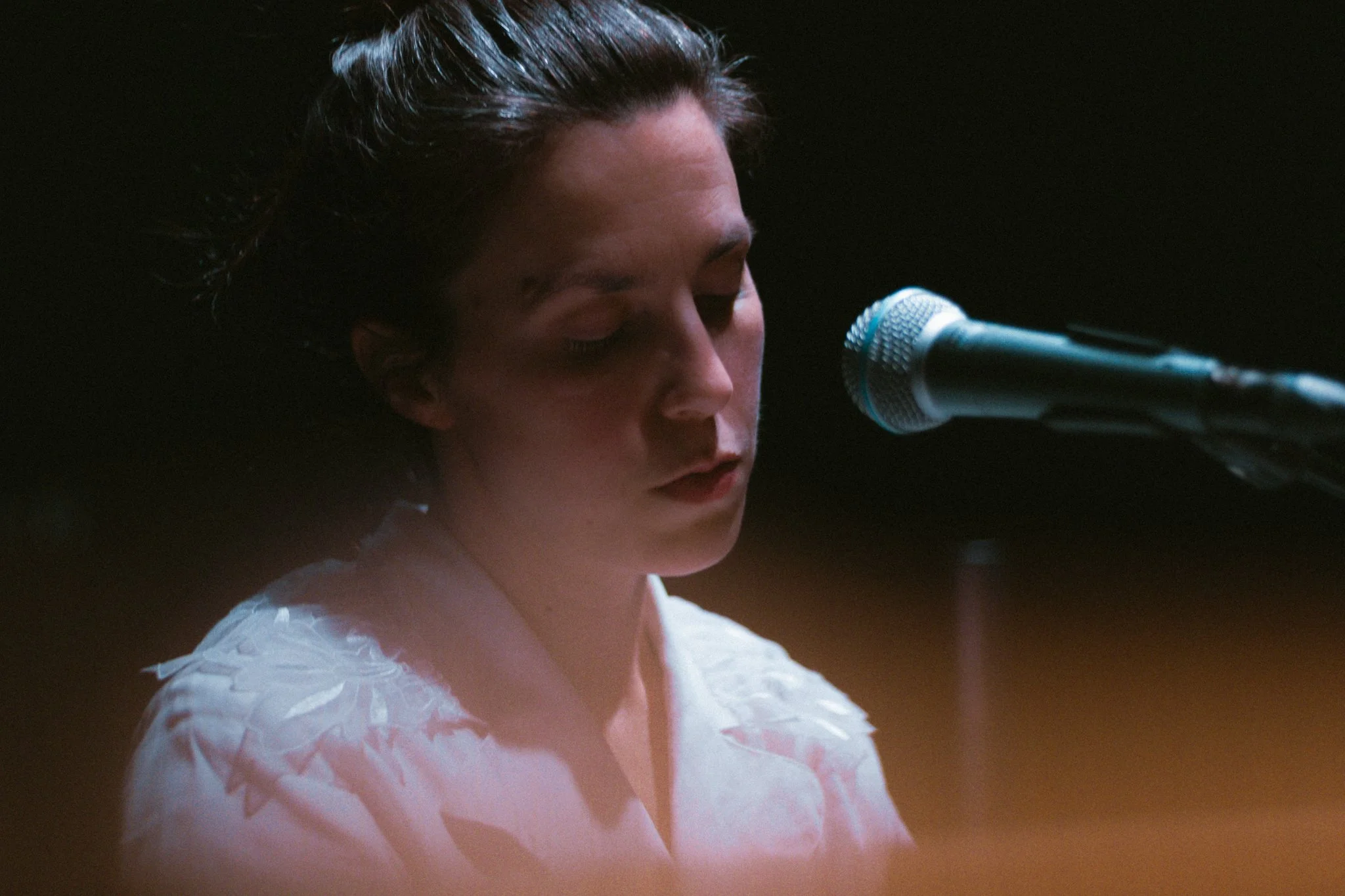 A woman with dark hair styled back, closed eyes, and a contemplative expression, sitting at a microphone in a dimly lit setting.