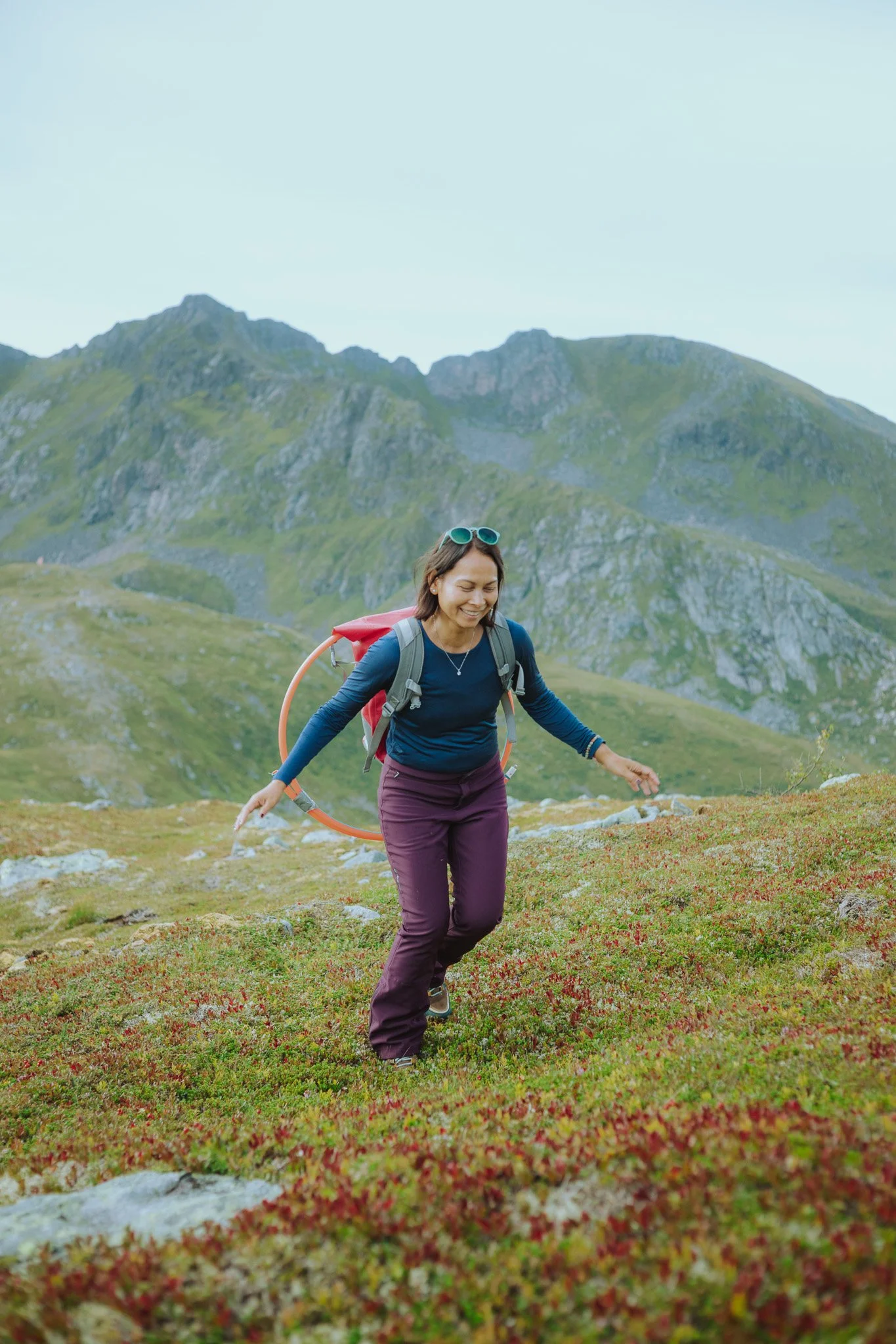 A woman hiking in a mountainous area with green hills and rocky peaks, wearing a blue long-sleeve shirt, purple pants, and carrying a red backpack.