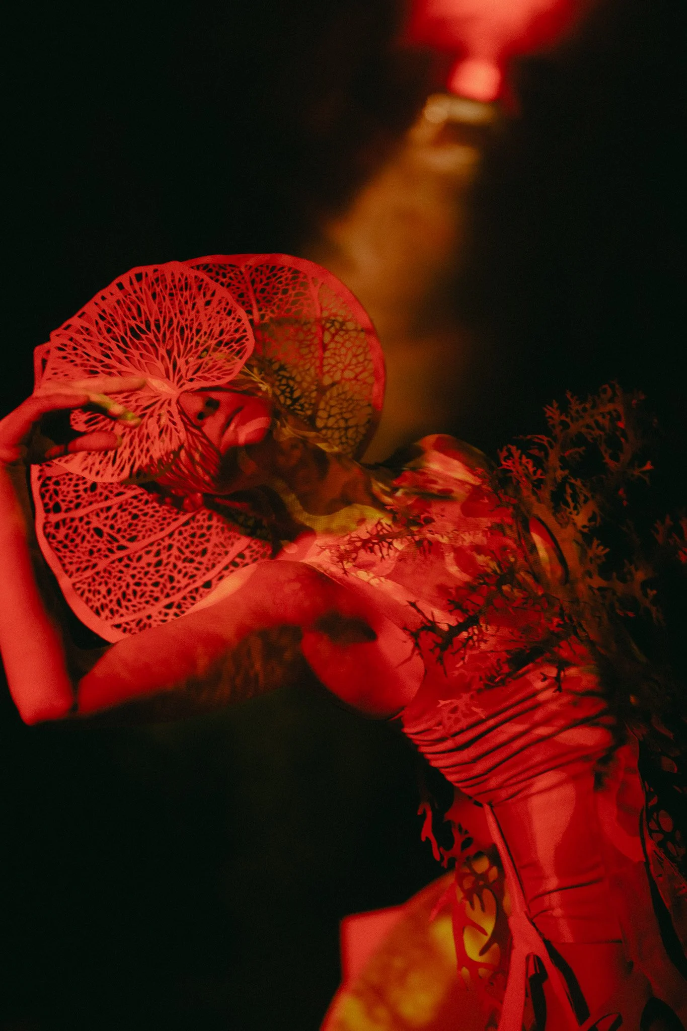 A model wearing intricate, alien-like red accessories and red clothing with a dark background, illuminated by red lighting.