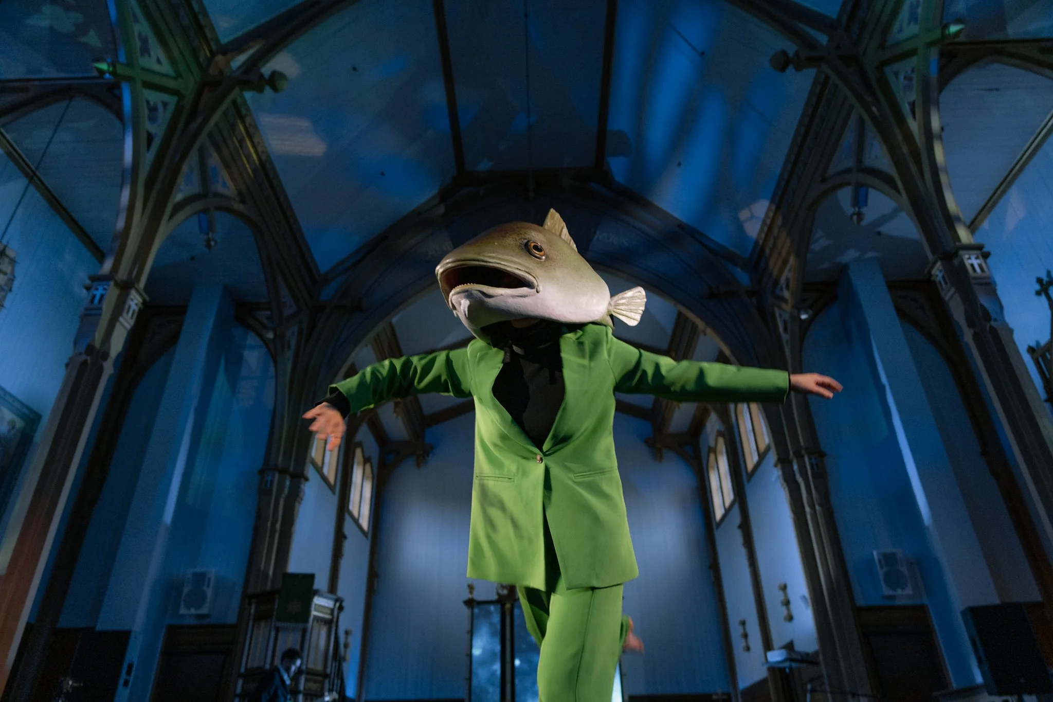 A person wearing a green suit with arms outstretched and a fish head mask, standing inside a church with arched ceiling and stained glass windows.