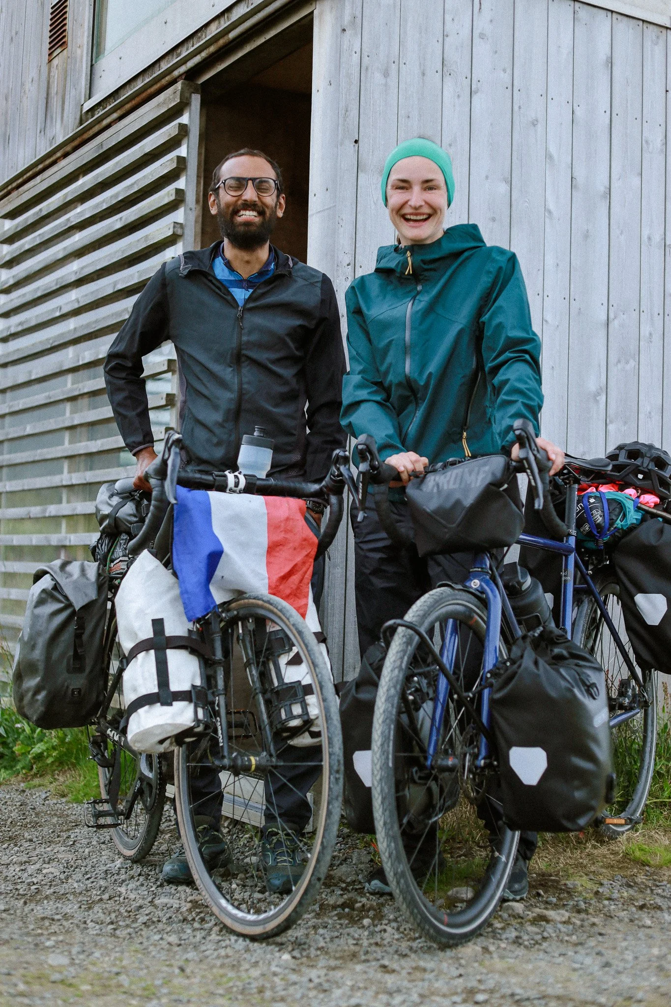 Two smiling cyclists posing with their loaded touring bikes in front of a wooden building.