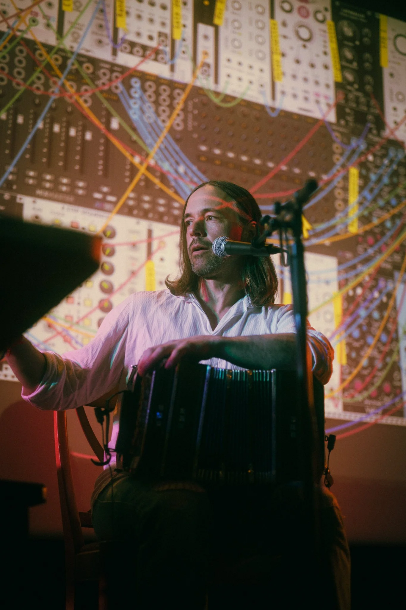 A man with long hair and a beard sitting in front of an electronic music setup, with a microphone and a percussion instrument, and a large digital visual display with complex patterns in the background.