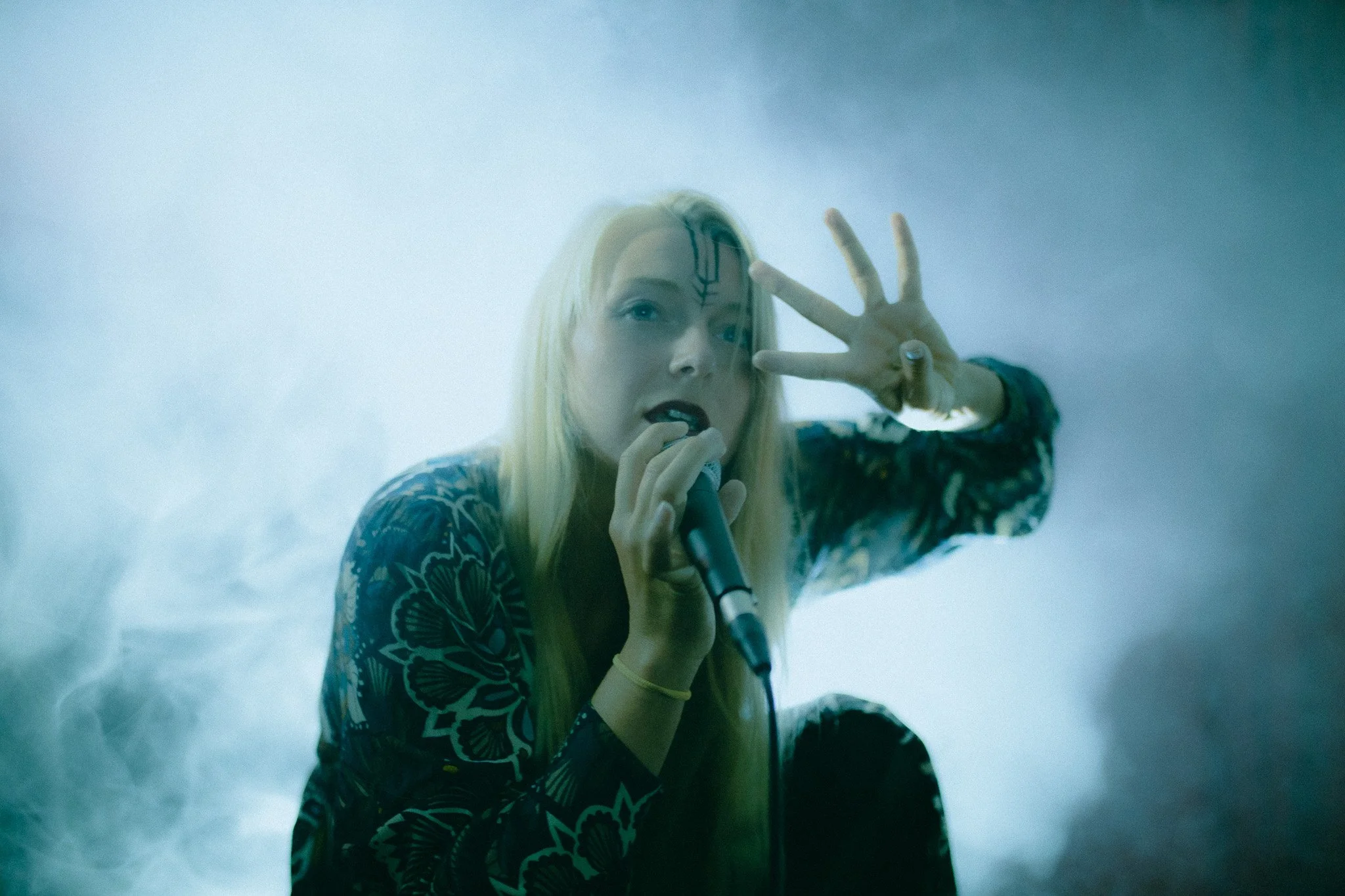 A woman with long blonde hair, dark lipstick, and makeup is holding a microphone close to her mouth and making a peace sign with her left hand. She has a decorative tattoo or face paint on her forehead and is wearing a dark, patterned top. The scene 