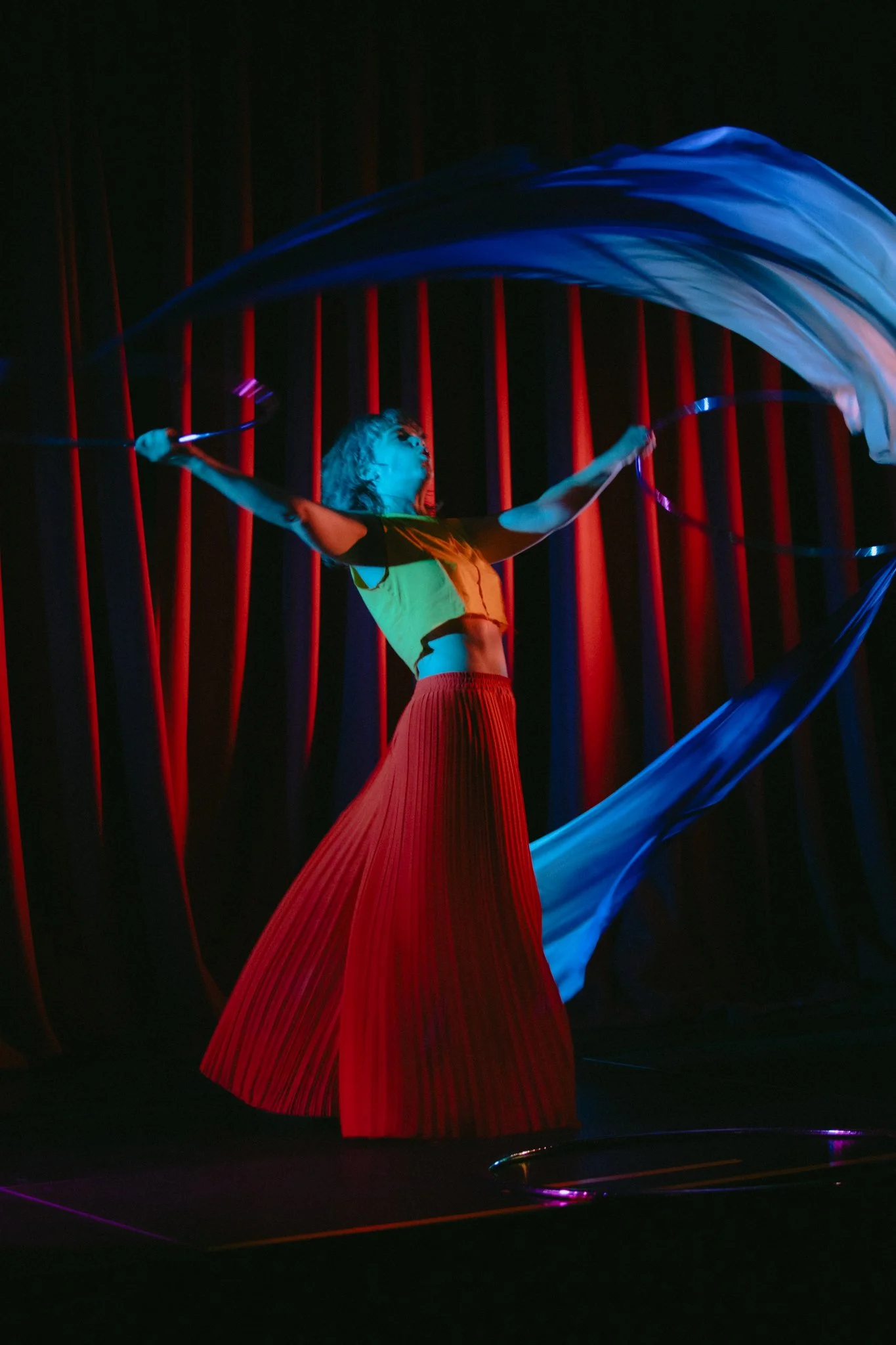 A performer on stage, illuminated by colorful lights, waving blue and white cloths with red vertical stripes in the background.