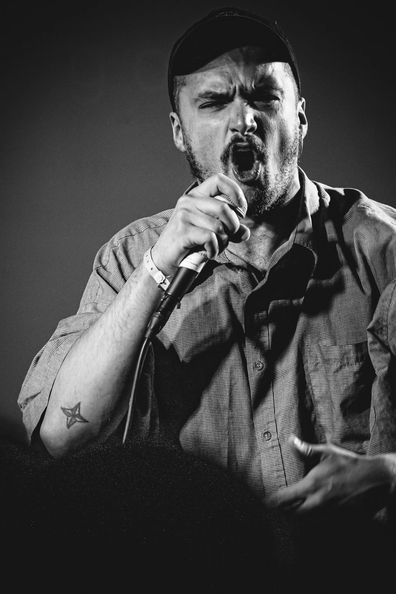 A man singing passionately into a microphone, wearing a baseball cap and a short-sleeved button-up shirt, with a star tattoo on his left forearm.