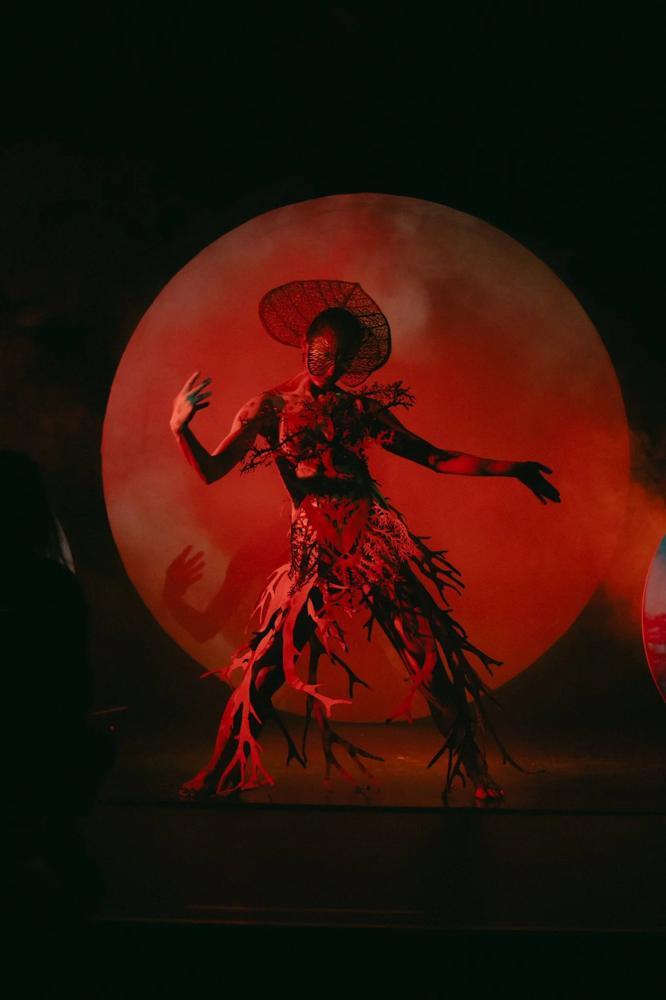 Performer dressed in a nature-inspired costume with branches and leaves, dancing in front of a large, red moon.