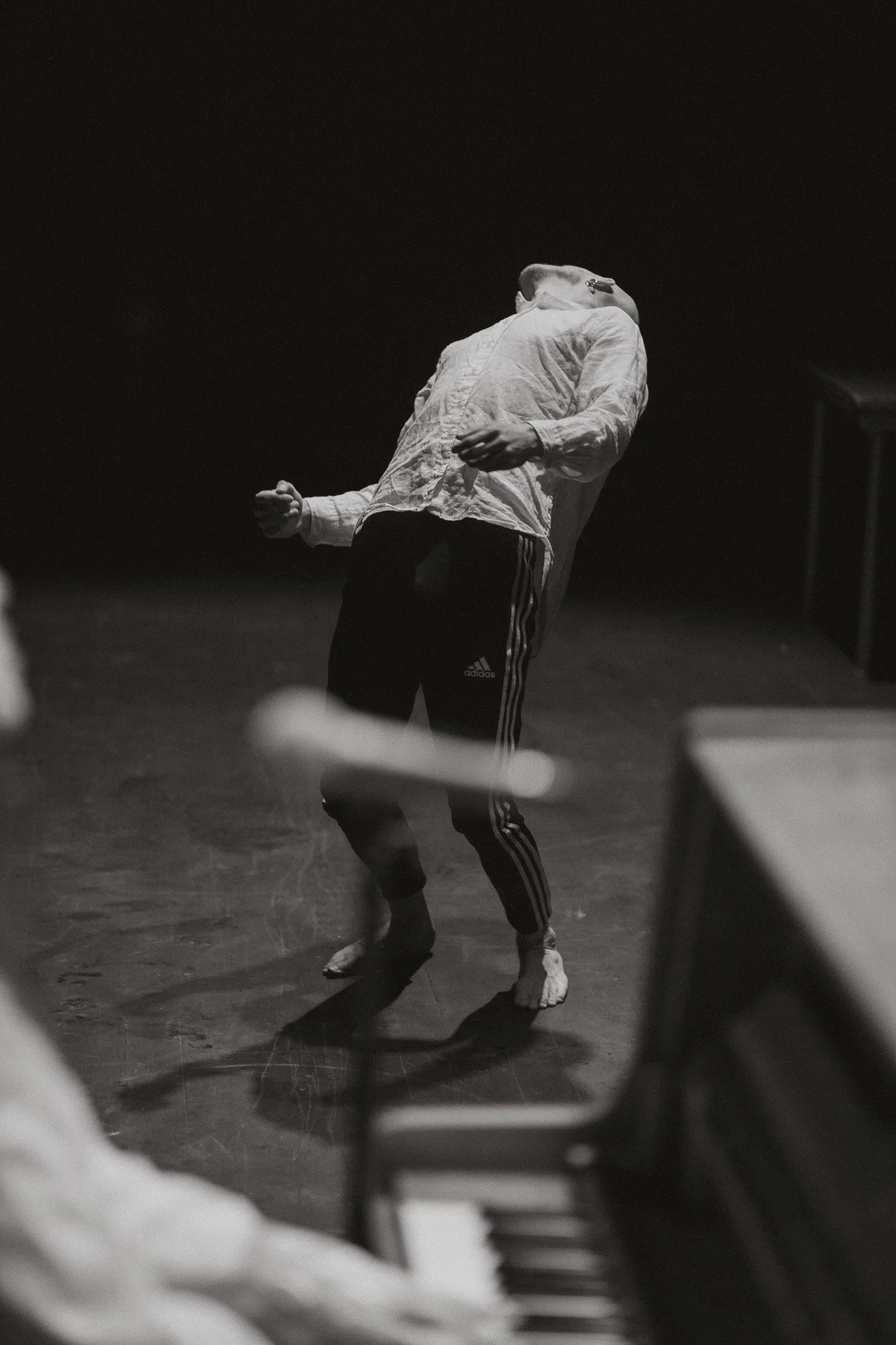 A person dancing barefoot on a dark stage, wearing a light-colored shirt and Adidas track pants, with their head tilted back and arms in expressive motion.
