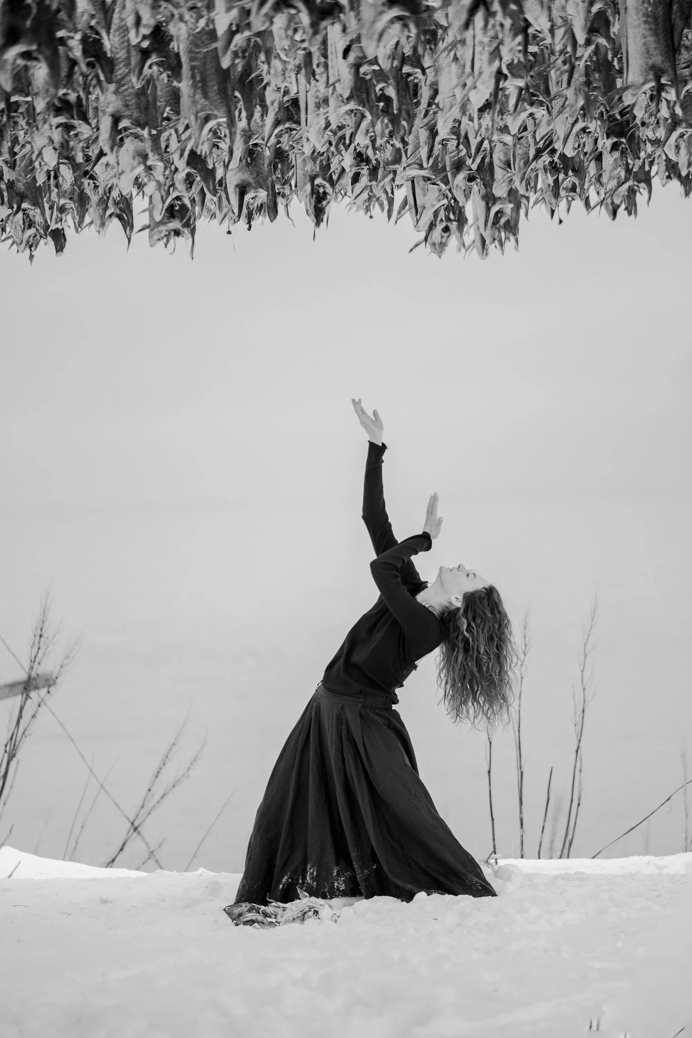 A woman in a black long-sleeve top and long skirt is kneeling in the snow outdoors, with her head tilted back and arms raised, appearing to dance or reach upward.