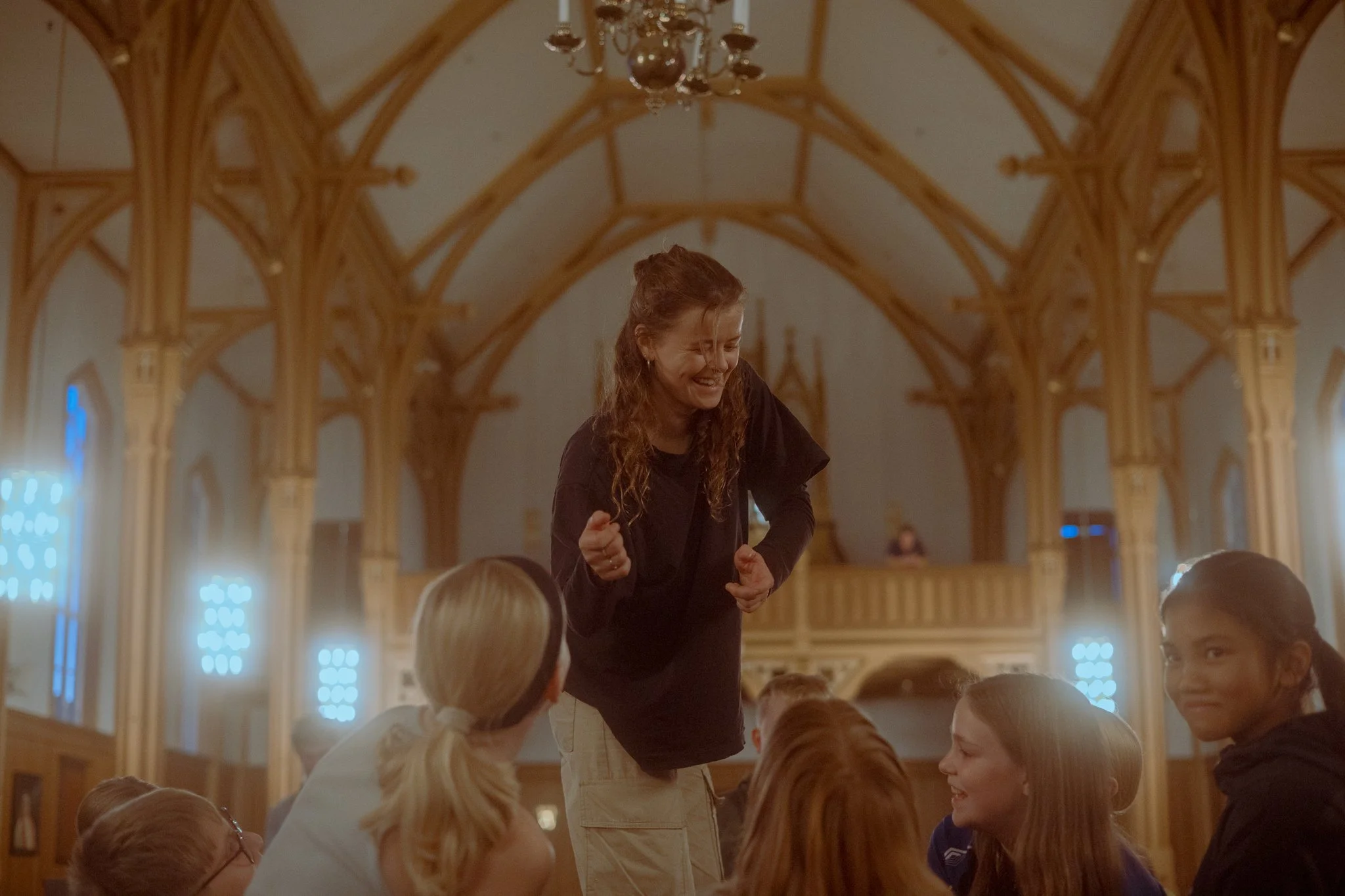 A woman with long curly hair, wearing a black shirt and beige pants, stands on a table, smiling and gesturing with clenched fists, surrounded by children sitting on the floor inside a church with wooden arches.