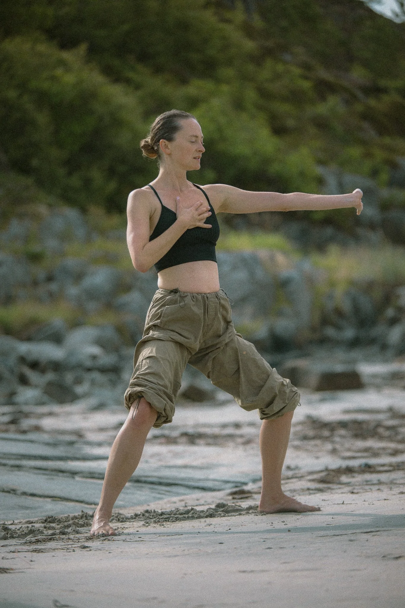 Woman practicing yoga outdoors on a sandy beach, doing a pose with one hand on her chest and the other extended outward.
