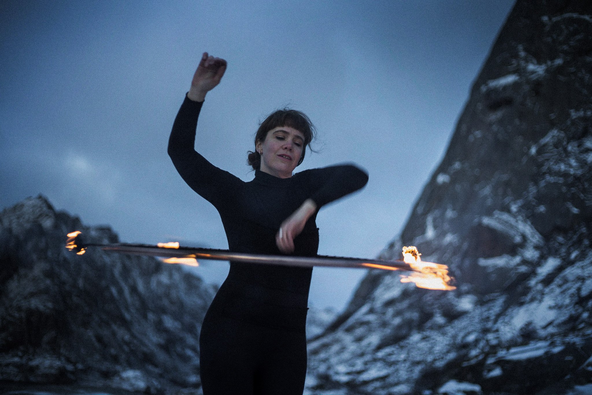 A woman in a black outfit spinning a flaming staff in a rocky outdoor area during twilight.