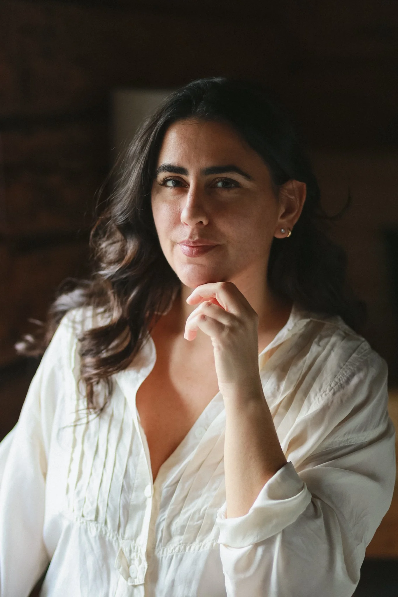 A woman with long dark wavy hair, wearing a white blouse, standing indoors with a wooden background, looking at the camera with a slight smile and resting her chin on her hand.